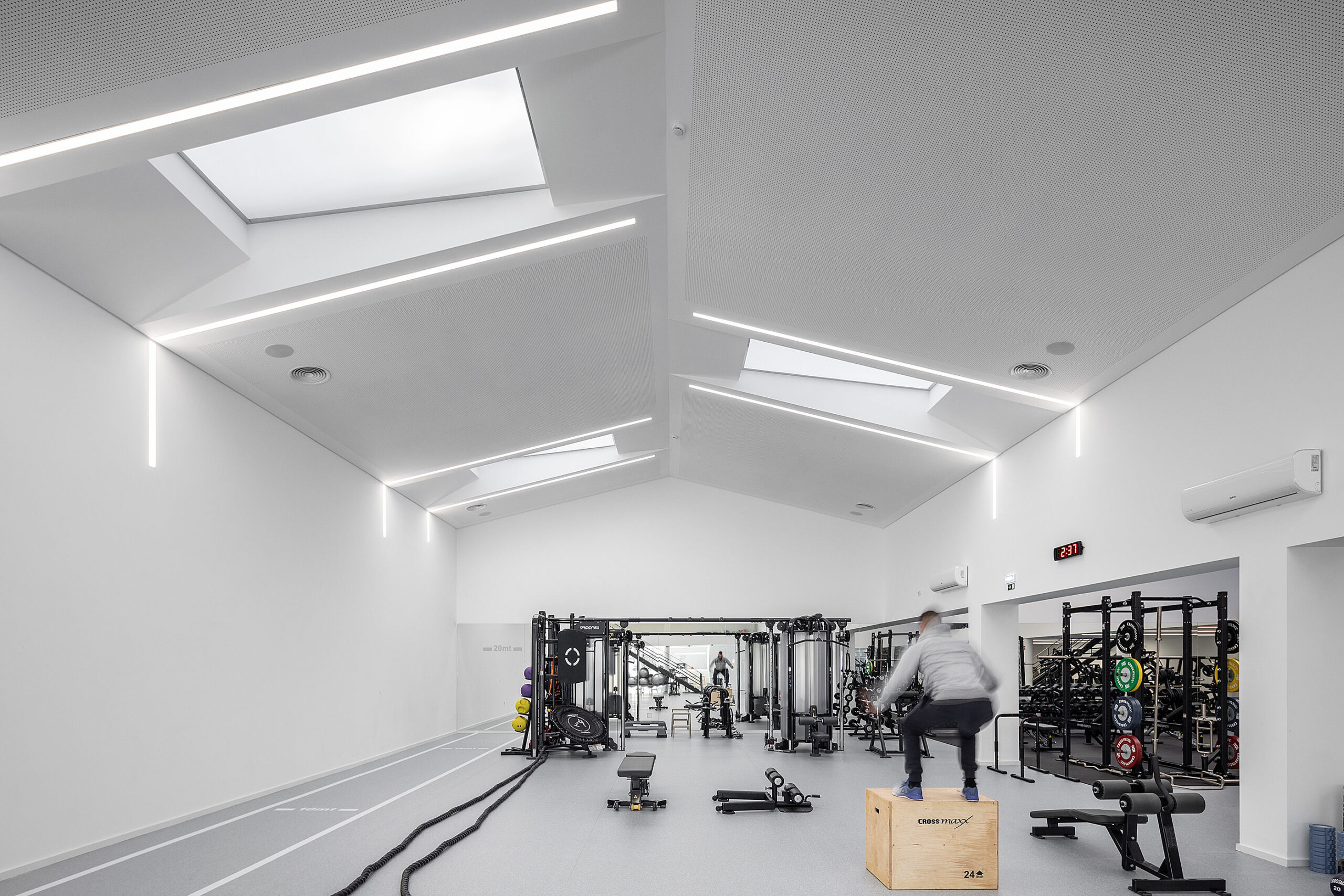 Ginásio com equipamentos, piso cinza com pista e claraboias. Iluminação linear e design minimalista. — Academia20 arquitetura StudioWorkers em Espinho