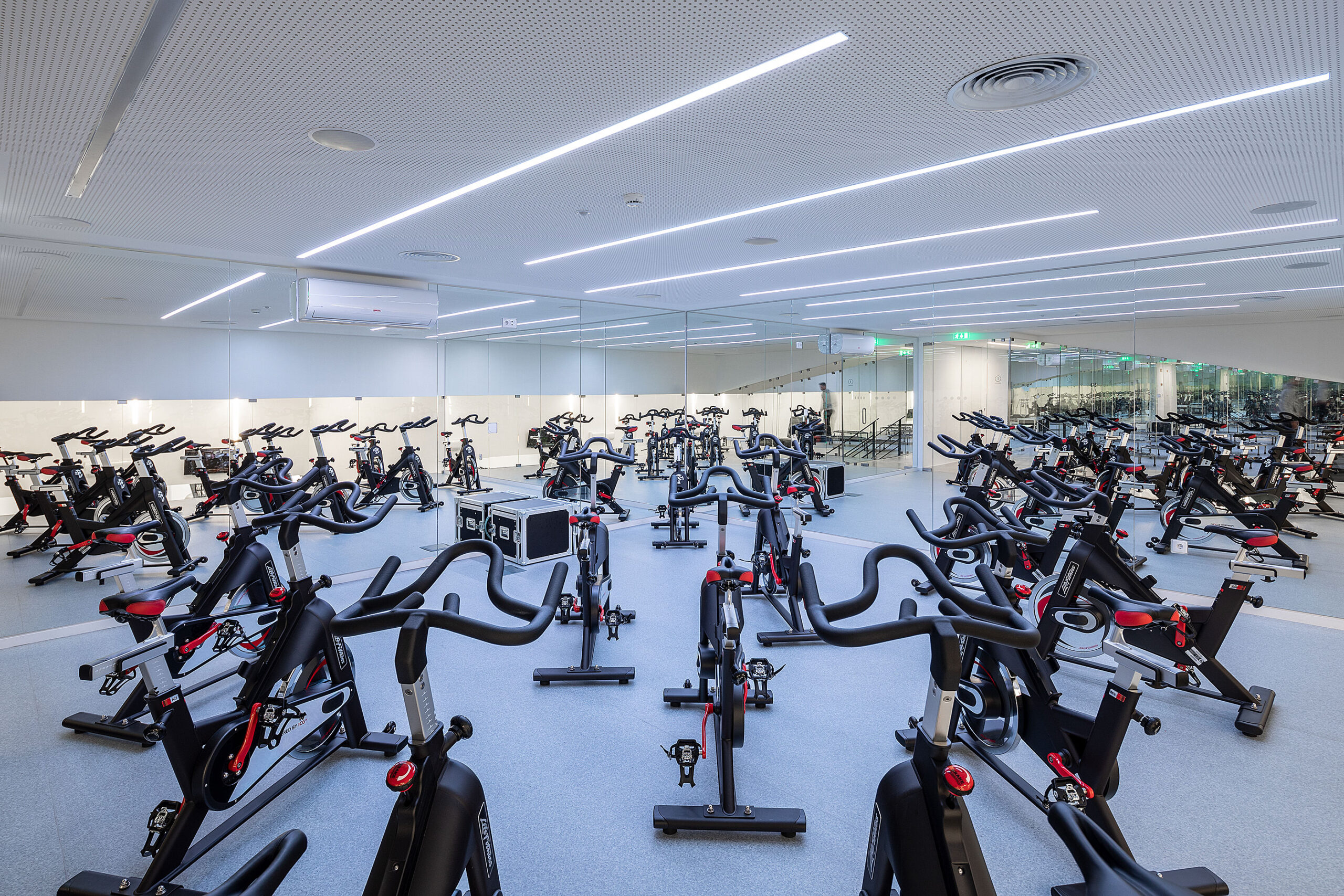 Sala de spinning com bicicletas pretas e vermelhas. Espelhos ampliam o espaço. Iluminação linear no teto. — Academia20 arquitetura StudioWorkers em Espinho