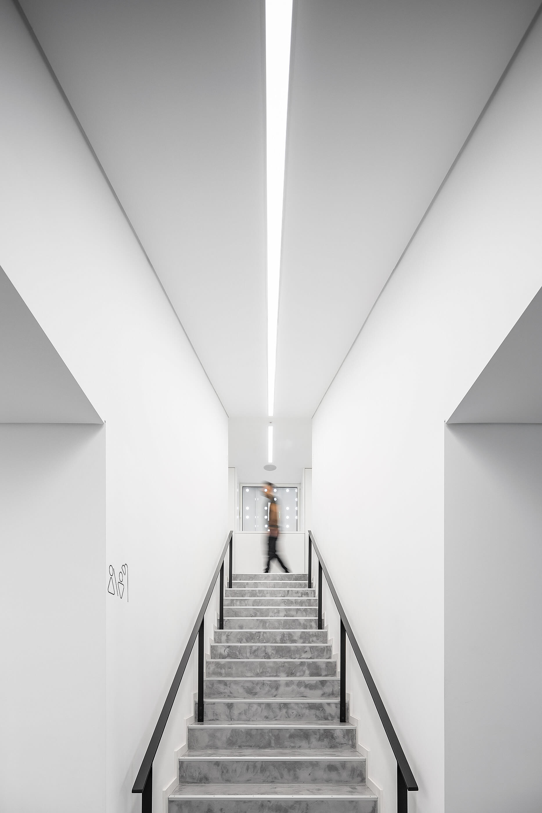 Escadas de betão com corrimão preto. Paredes brancas e iluminação linear. Pessoa a subir. — Academia20 arquitetura StudioWorkers em Espinho