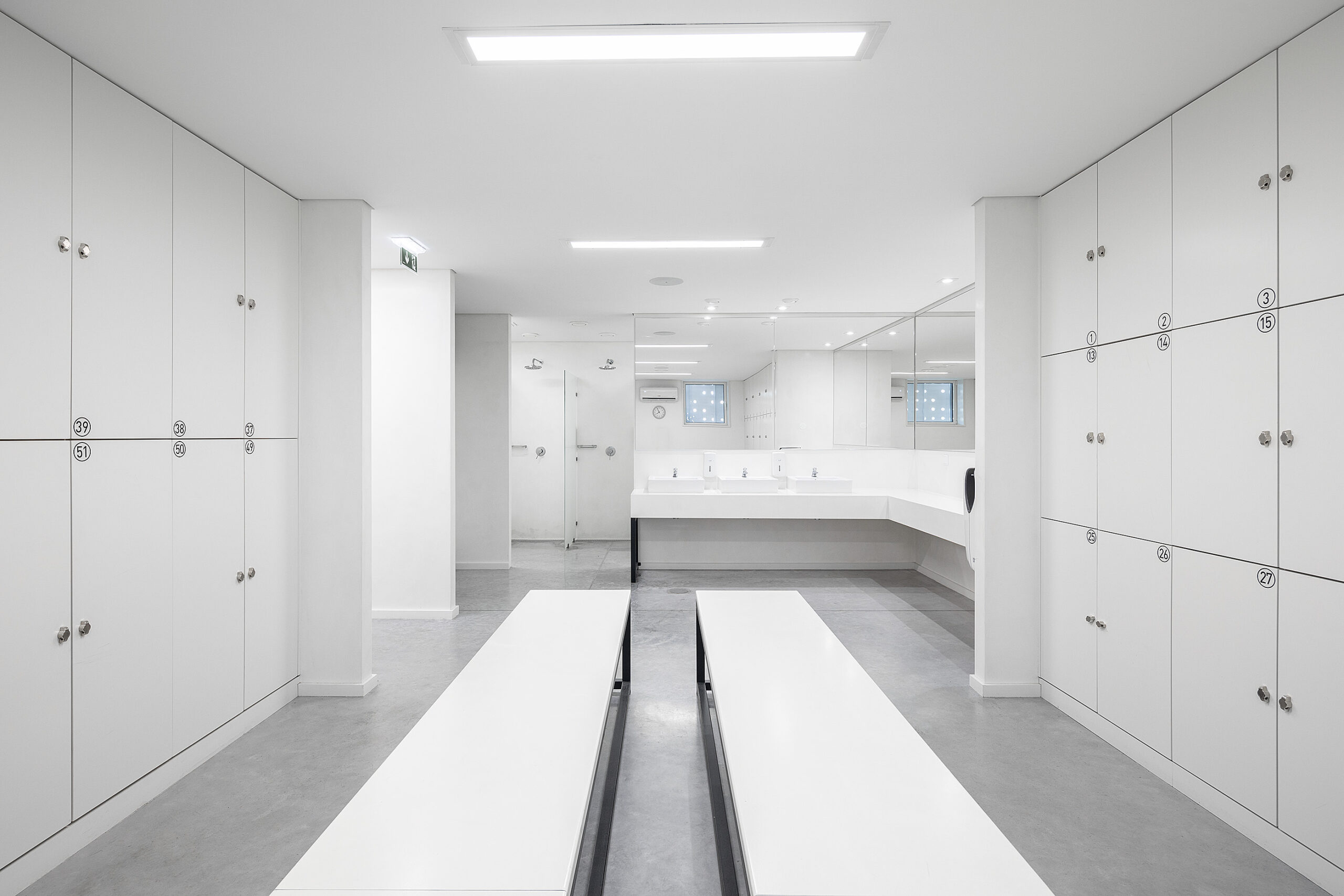 Vestiário amplo com cacifos brancos, lavatórios e espelhos. Bancos centrais e iluminação linear. — Academia20 arquitetura StudioWorkers em Espinho