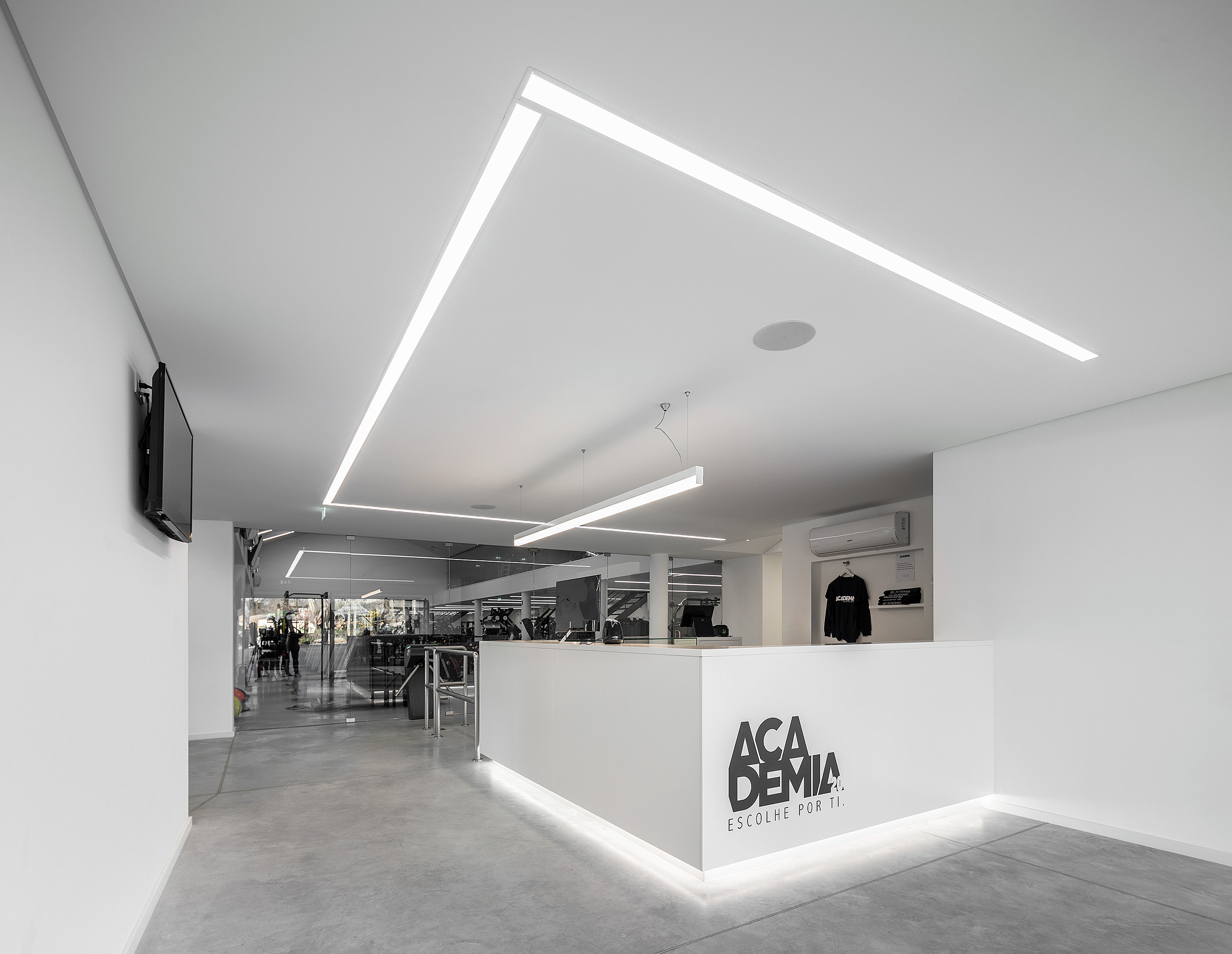 Recepção com balcão branco iluminado e logótipo. Piso cinza e vista para o ginásio. — Academia20 arquitetura StudioWorkers em Espinho