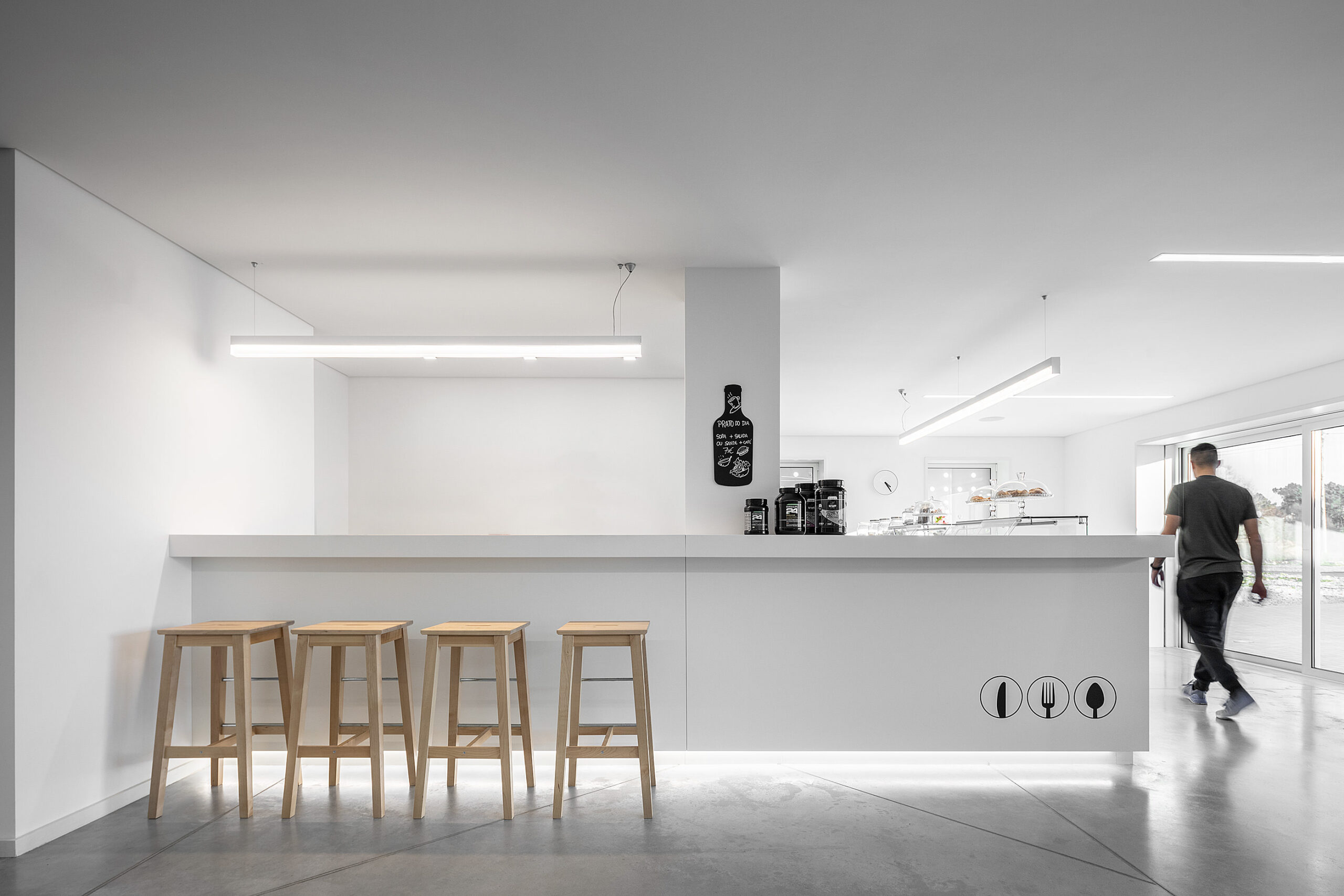 Cafetaria com balcão branco, bancos de madeira e iluminação linear. Piso cinza e design minimalista. — Academia20 arquitetura StudioWorkers em Espinho