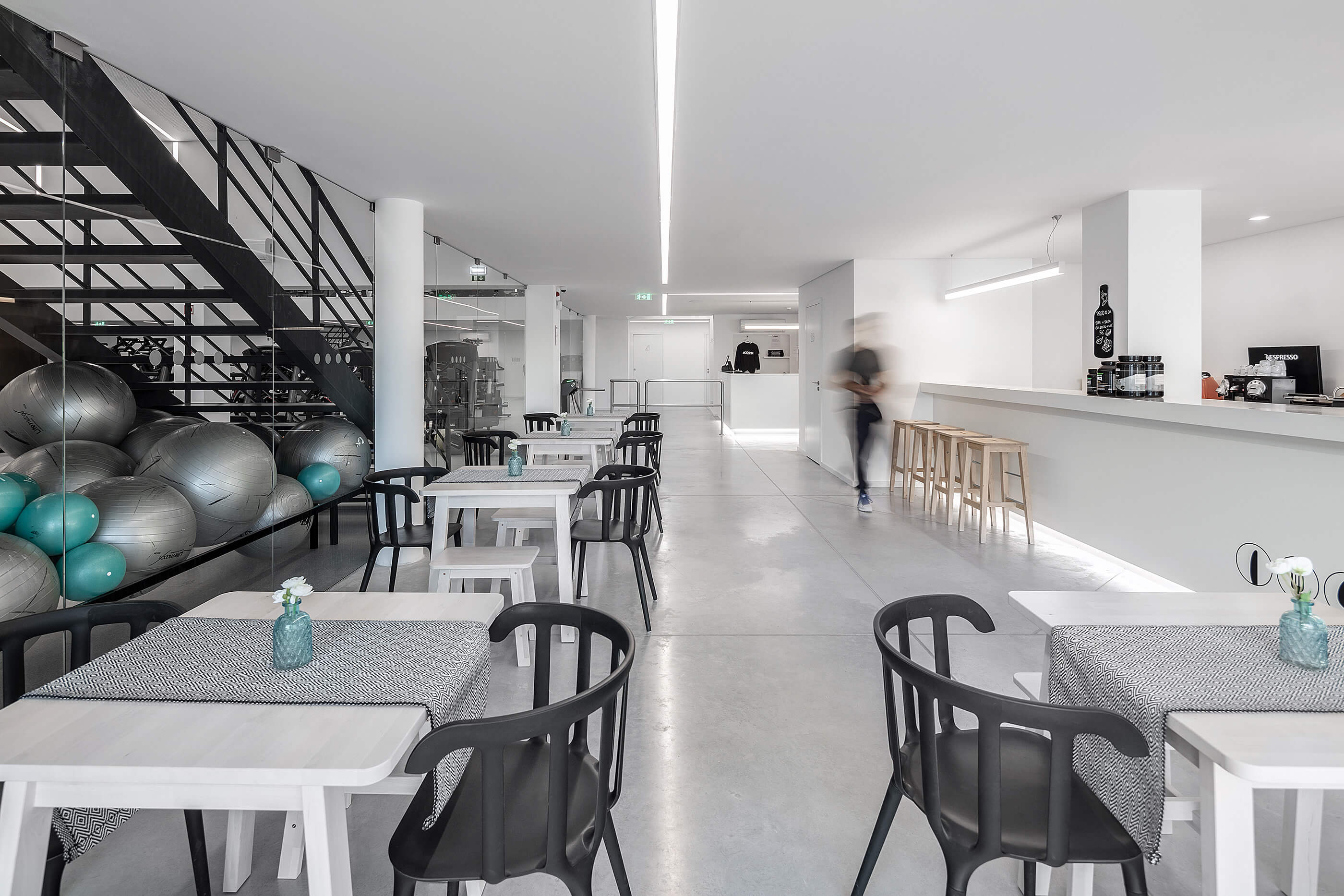 Cafetaria com mesas e cadeiras pretas e brancas. Escada metálica e vista para o ginásio. — Academia20 arquitetura StudioWorkers em Espinho