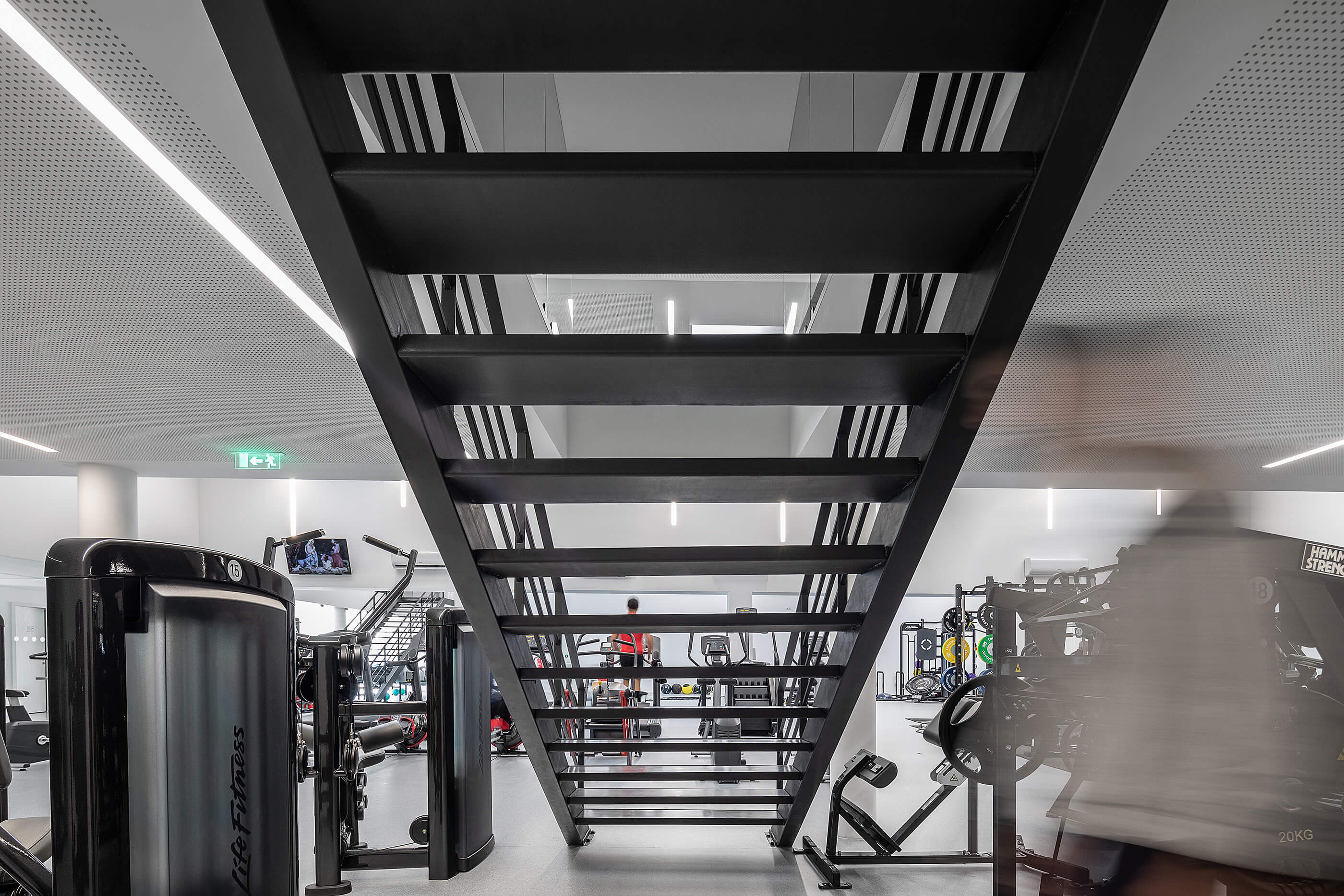 Ginásio com vista para escada metálica preta. Equipamentos de exercício e iluminação linear no teto branco. — Academia20 arquitetura StudioWorkers em Espinho