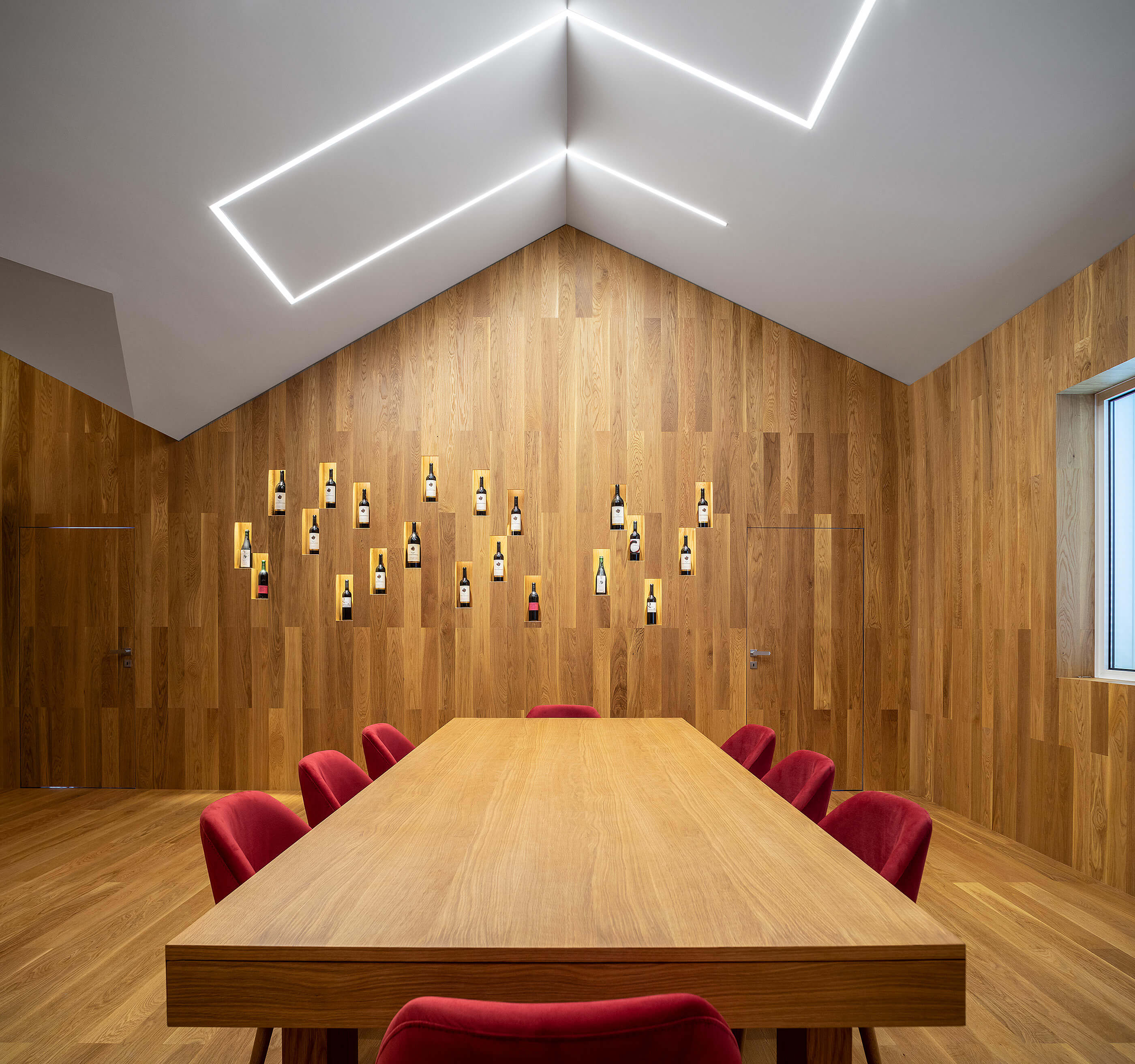 Sala de provas com mesa de madeira, cadeiras vermelhas e parede de madeira com garrafas. — Adega do Castanheirinho arquitetura João Albano Arquitecto em Águeda
