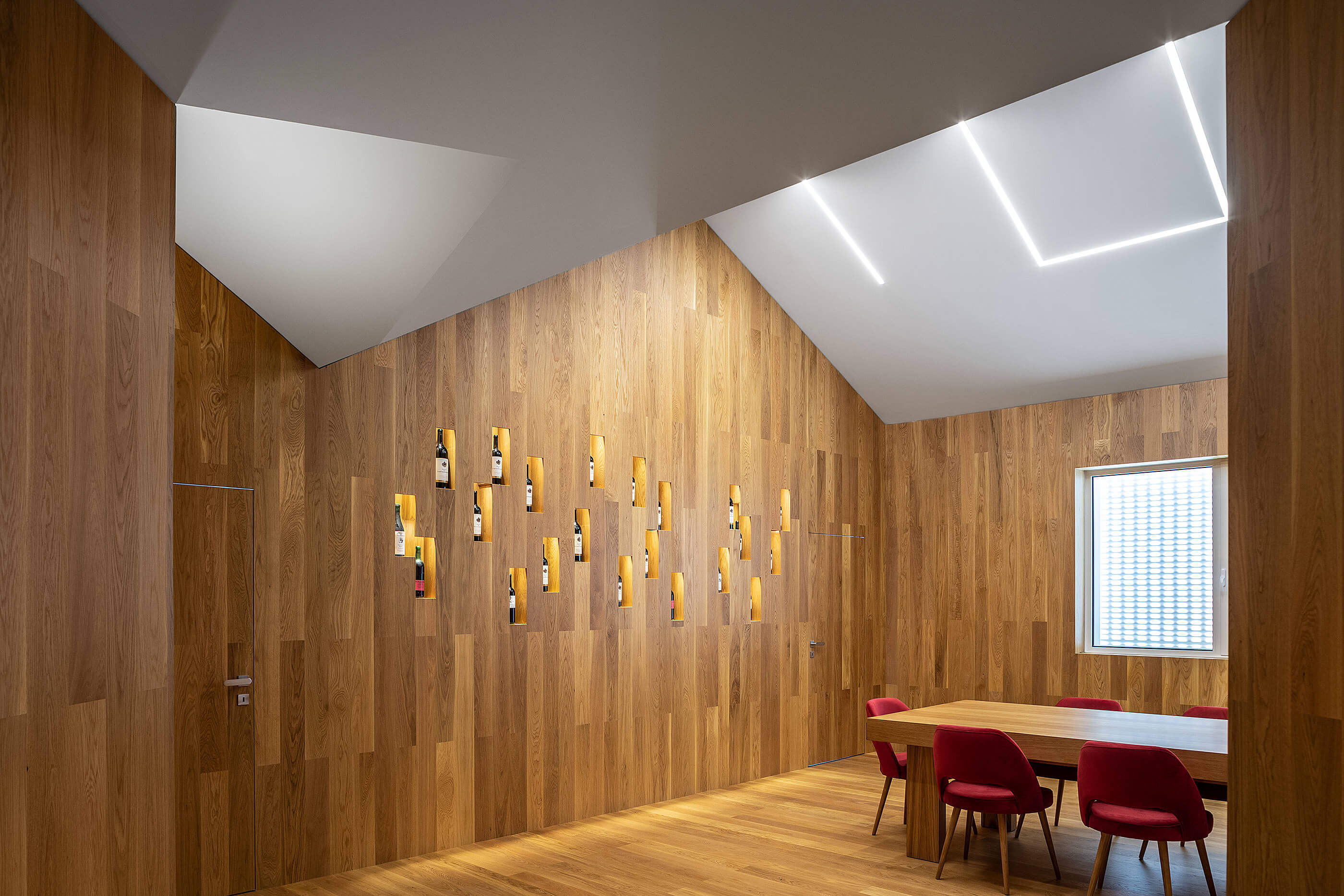 Sala de provas com mesa de madeira, cadeiras vermelhas e parede de madeira com garrafas. — Adega do Castanheirinho arquitetura João Albano Arquitecto em Águeda