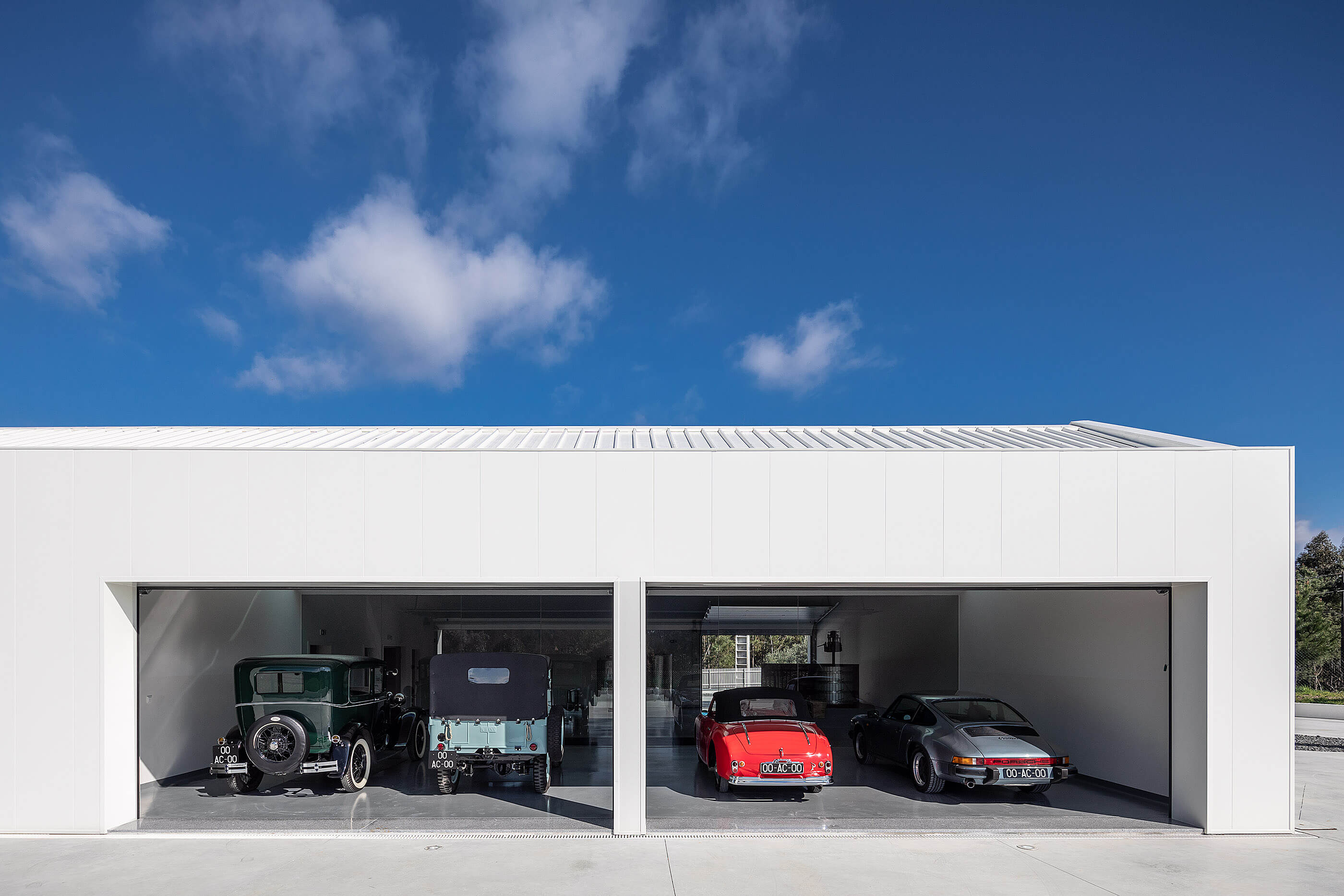 Garagem branca minimalista com dois portões abertos, exibindo carros clássicos sob céu azul. — Adega do Castanheirinho arquitetura João Albano Arquitecto em Águeda