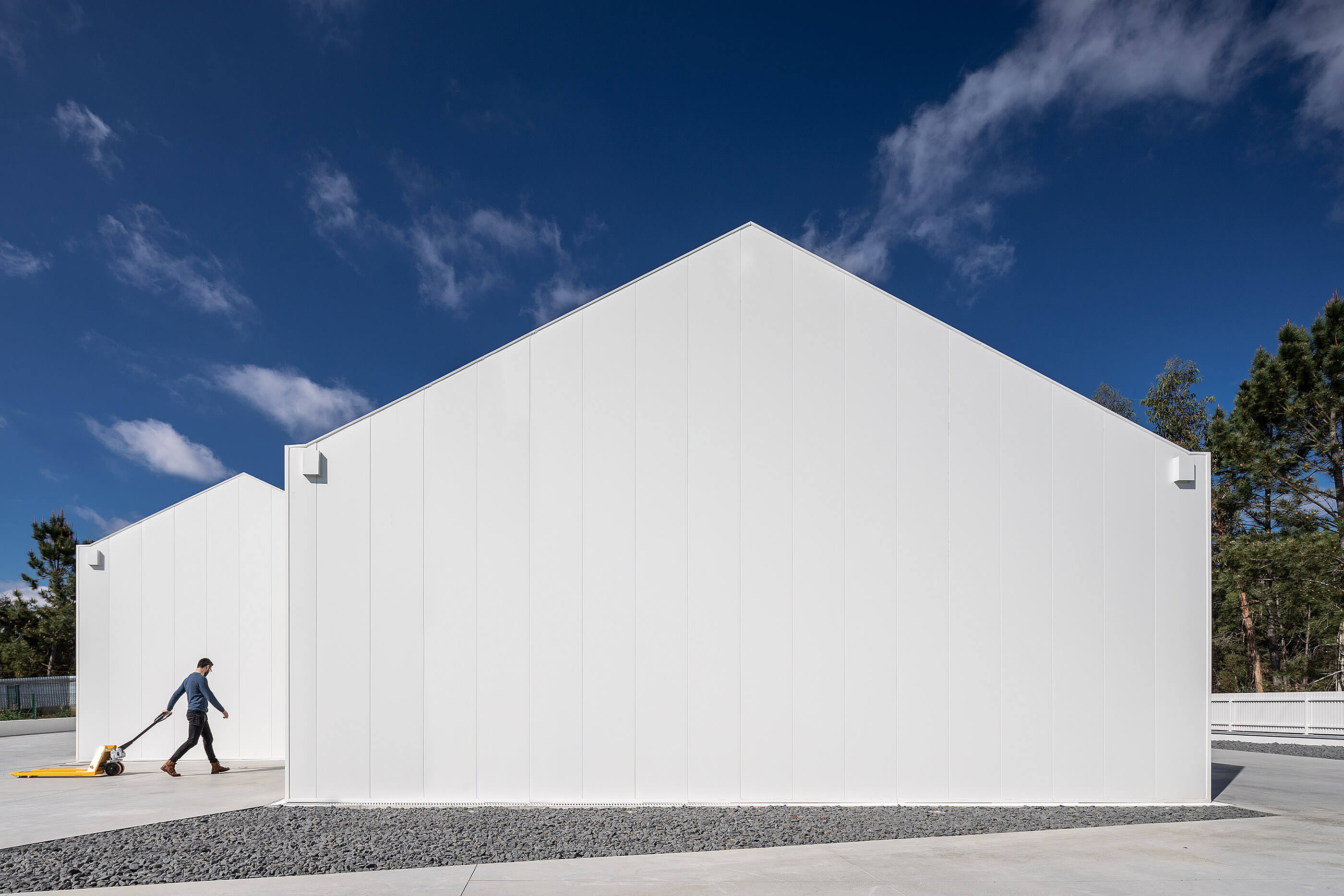Volume branco da adega com telhado em duas águas, homem a puxar carrinho manual. — Adega do Castanheirinho arquitetura João Albano Arquitecto em Águeda
