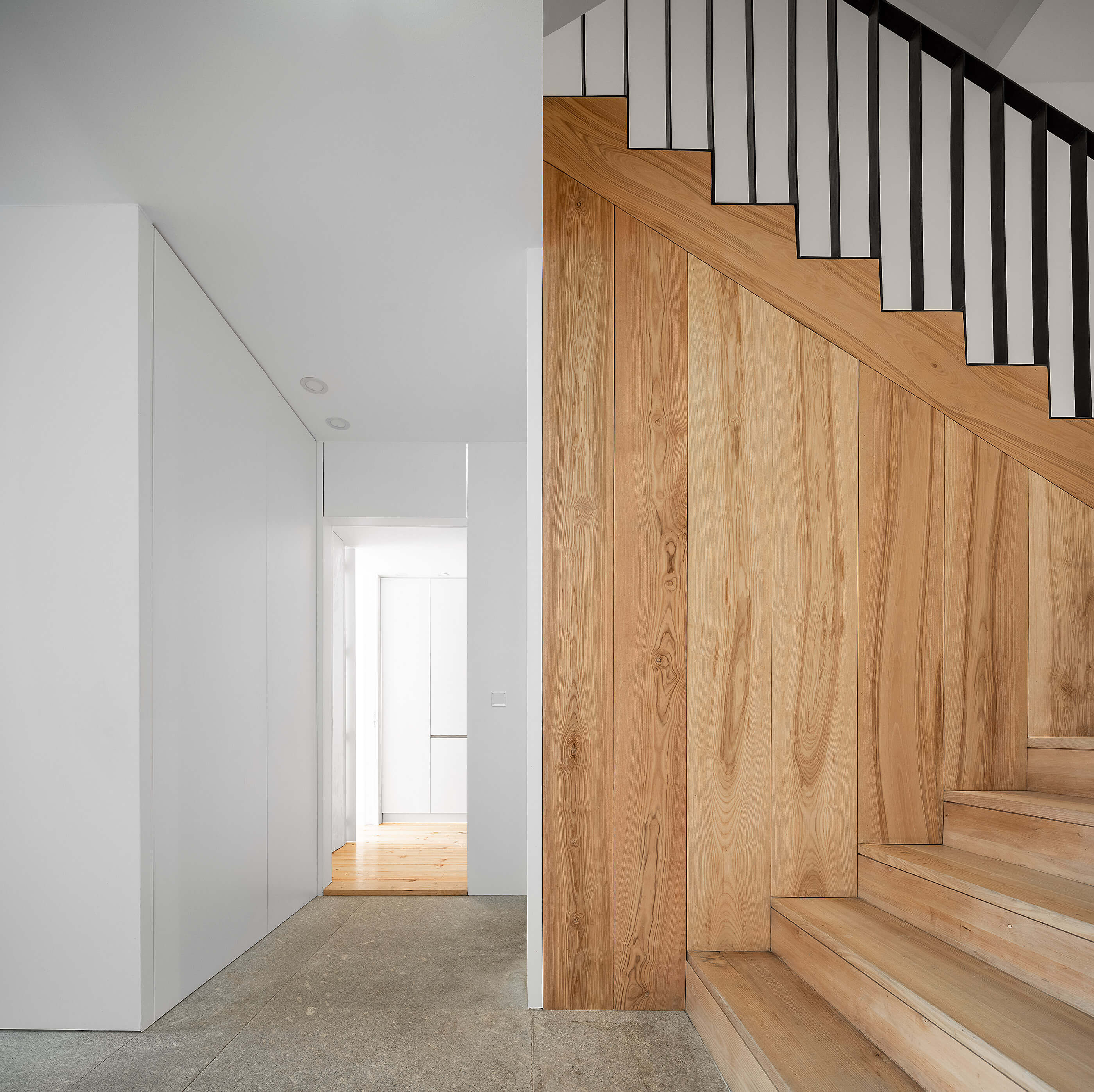 Hall de entrada com escadas de madeira clara, corrimão preto, paredes brancas e pavimento em pedra cinzenta. — Edifício Multifamiliar Alegria arquitetura MiMool Arquitectura & Interiores em Porto