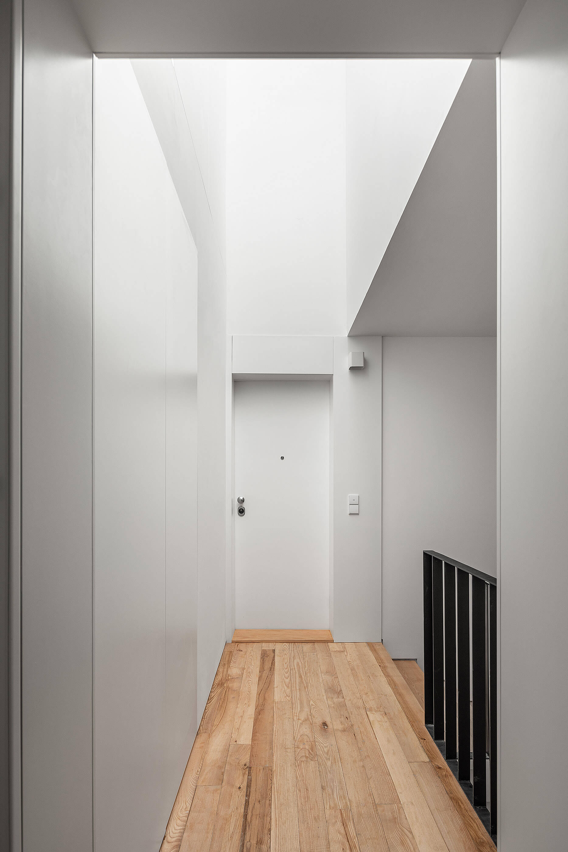 Corredor com chão em madeira clara, paredes brancas, porta branca e claraboia que ilumina o espaço. — Edifício Multifamiliar Alegria arquitetura MiMool Arquitectura & Interiores em Porto