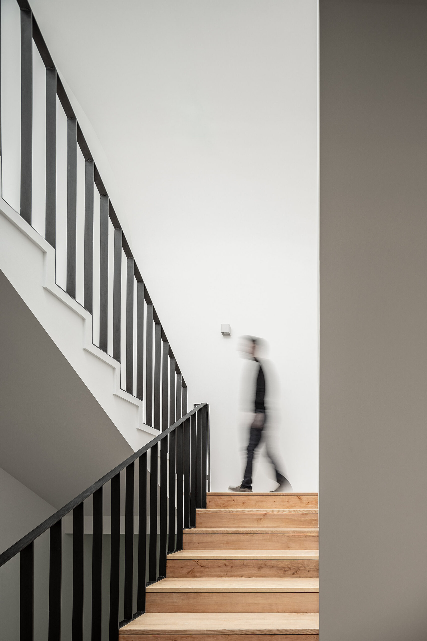 Escadas de madeira clara com corrimão preto, paredes brancas e pessoa desfocada a subir os degraus. — Edifício Multifamiliar Alegria arquitetura MiMool Arquitectura & Interiores em Porto