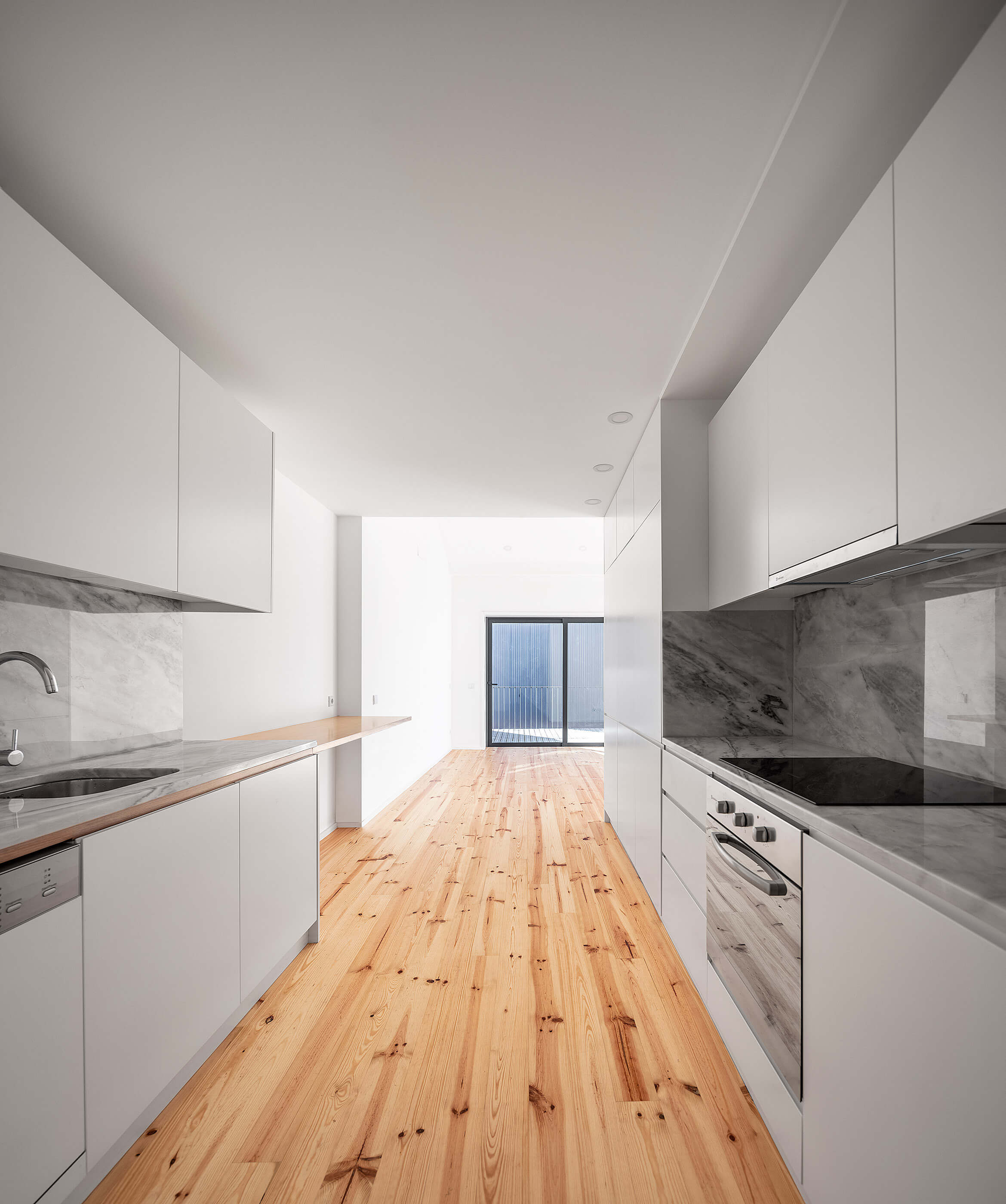 Cozinha comprida com armários brancos, bancada em mármore cinza, chão de madeira clara e acesso a varanda. — Edifício Multifamiliar Alegria arquitetura MiMool Arquitectura & Interiores em Porto