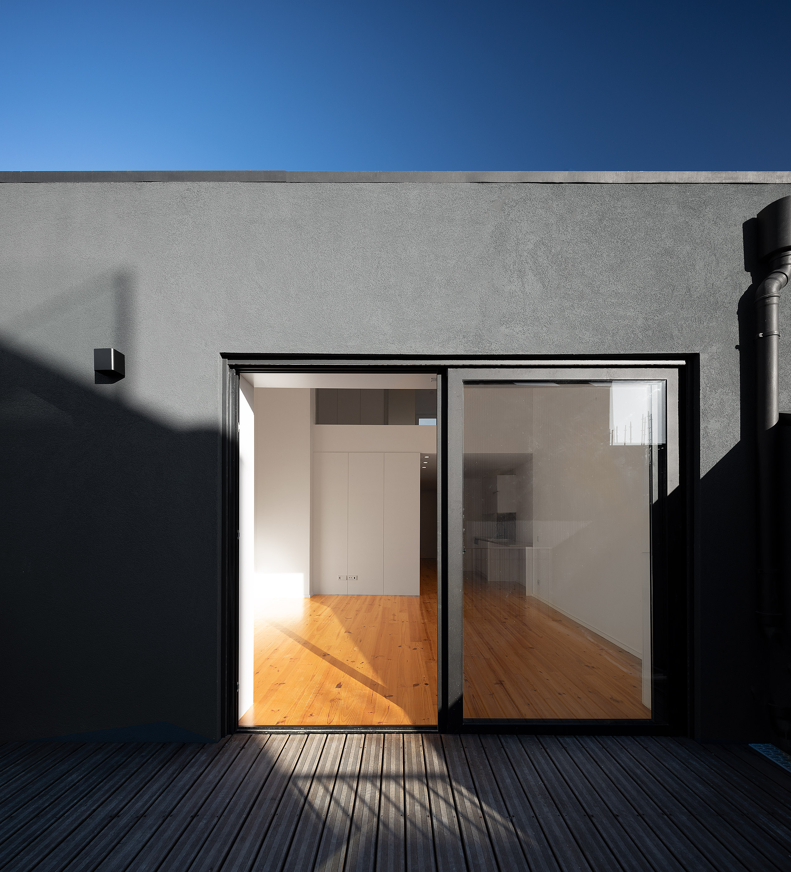 Terraço com deck de madeira escura, paredes cinzentas e porta de correr que dá acesso ao interior do apartamento. — Edifício Multifamiliar Alegria arquitetura MiMool Arquitectura & Interiores em Porto