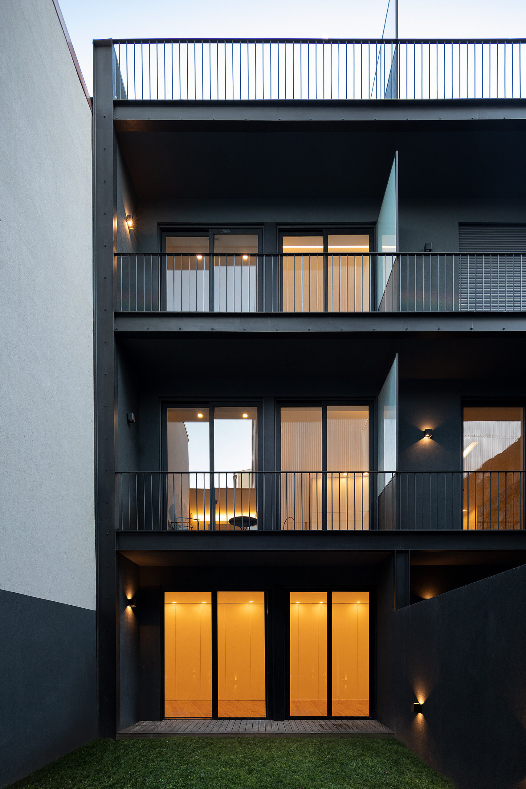 Fachada posterior do edifício com varandas, caixilharia preta e iluminação exterior. Jardim relvado em primeiro plano. — Edifício Multifamiliar Alegria arquitetura MiMool Arquitectura & Interiores em Porto