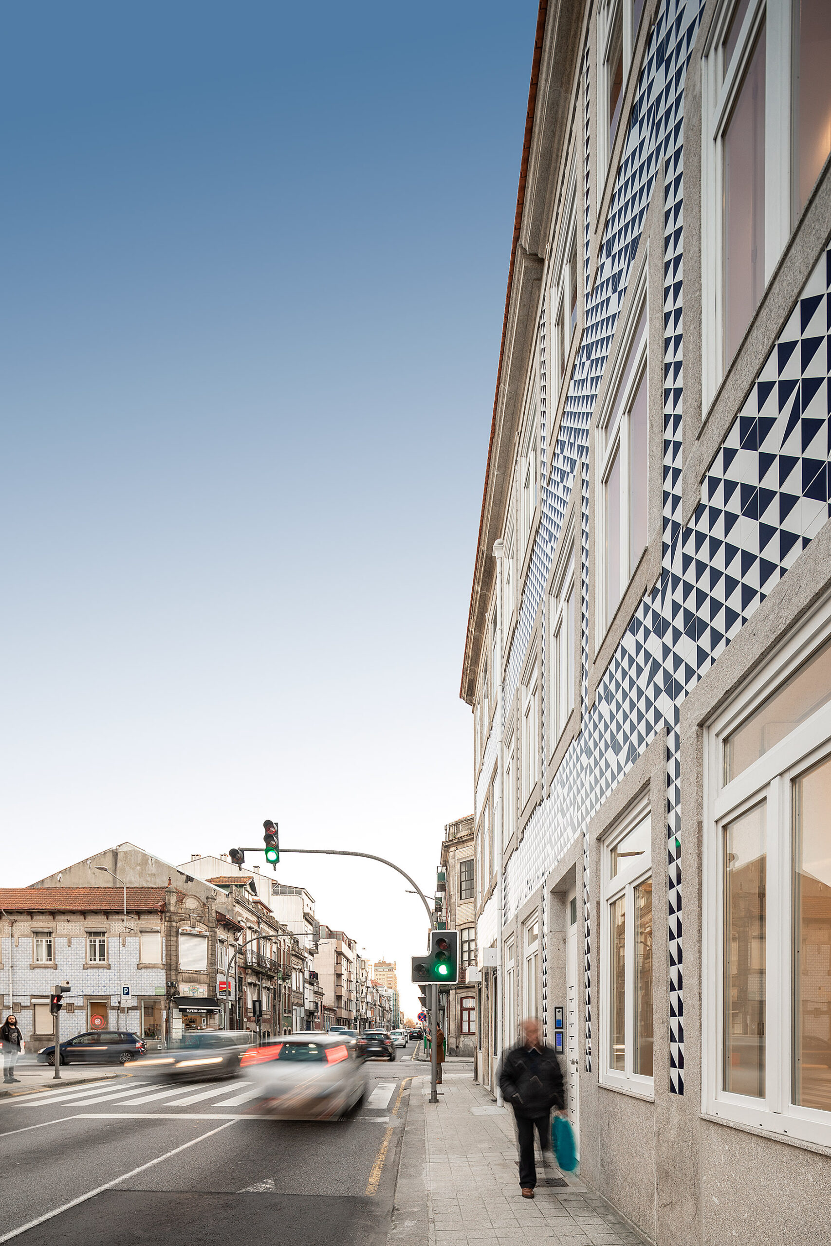 Edifício com fachada revestida a azulejos azuis e brancos, integrado na rua com trânsito e pessoas a circular. — Edifício Multifamiliar Alegria arquitetura MiMool Arquitectura & Interiores em Porto