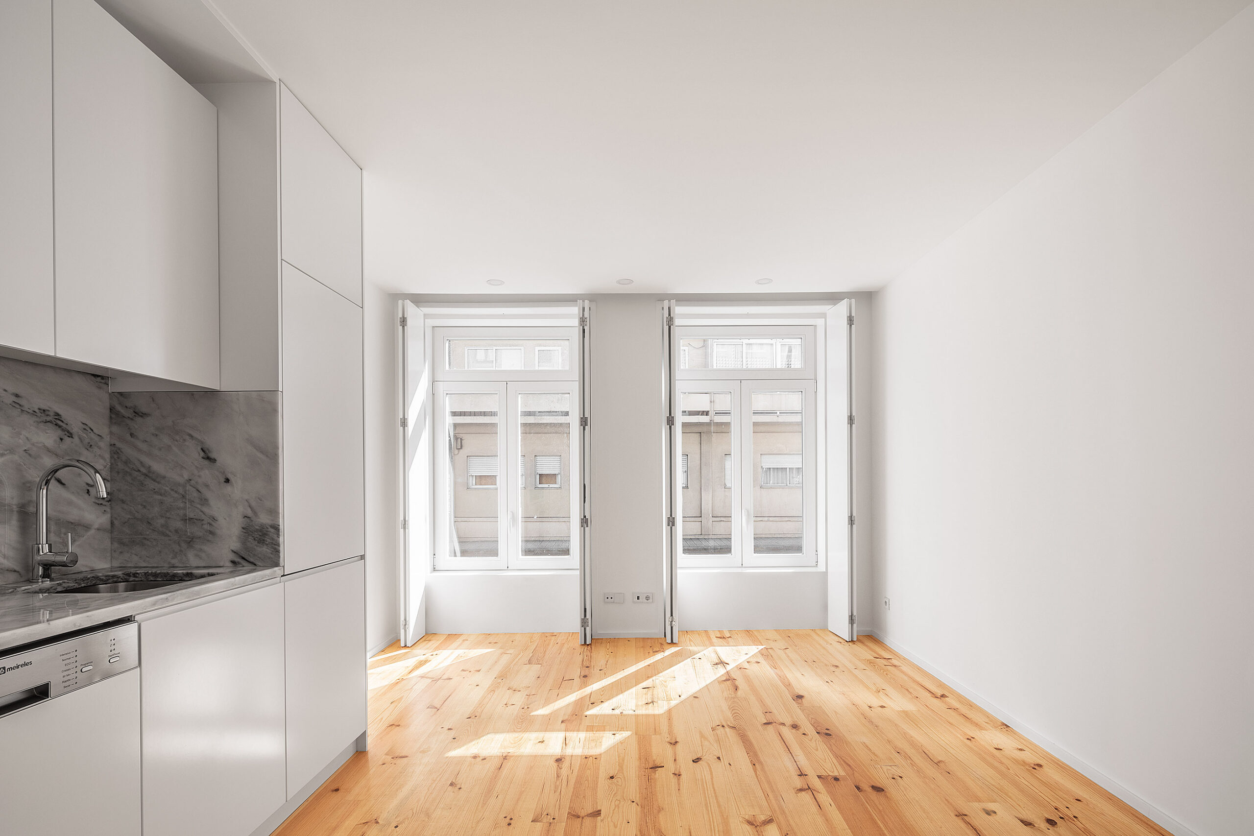 Cozinha integrada com armários brancos, bancada em mármore cinza, chão de madeira clara e janelas com portadas. — Edifício Multifamiliar Alegria arquitetura MiMool Arquitectura & Interiores em Porto