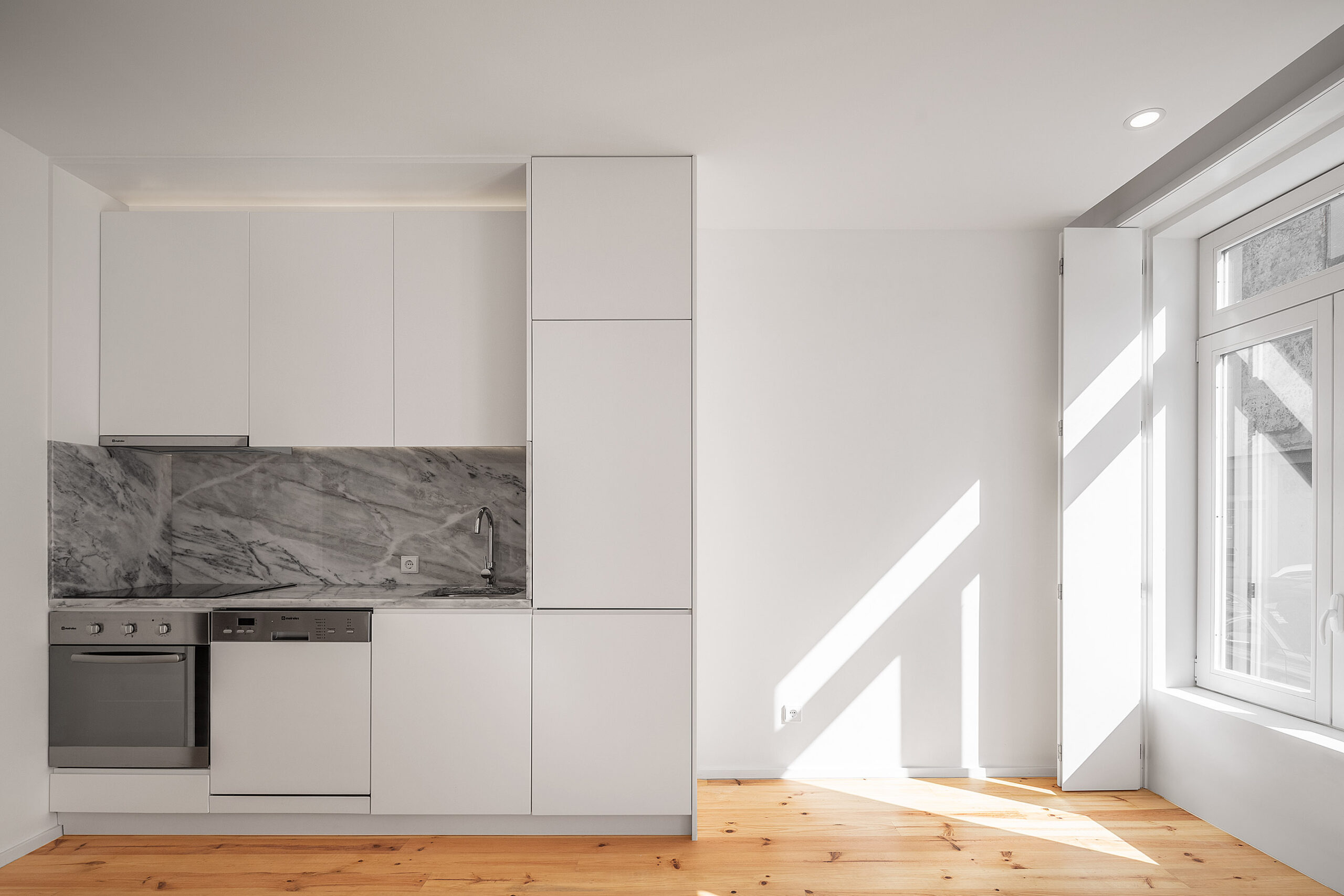 Cozinha moderna com armários brancos, bancada em mármore cinza, eletrodomésticos em inox e chão de madeira clara. — Edifício Multifamiliar Alegria arquitetura MiMool Arquitectura & Interiores em Porto