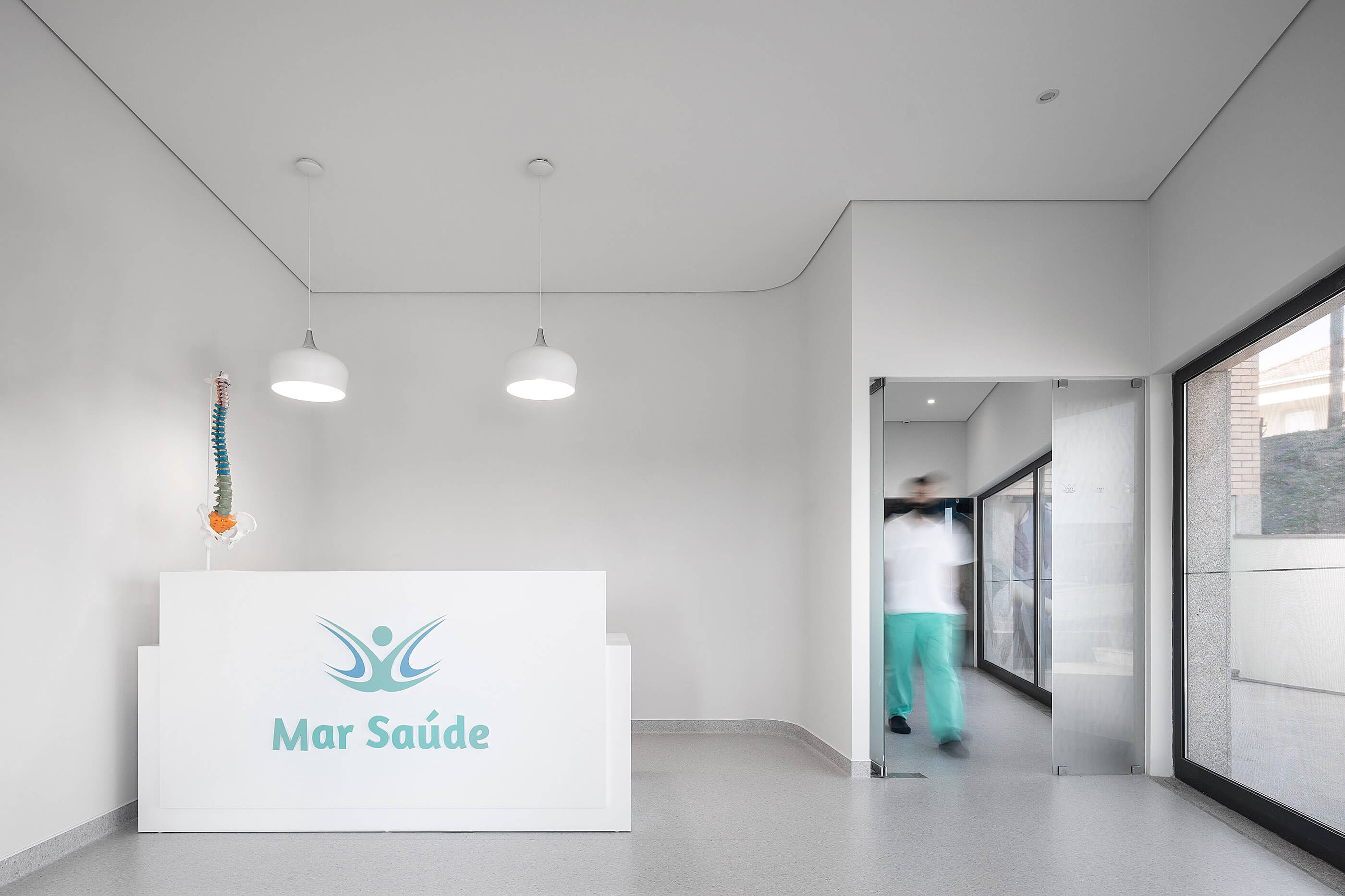 Recepção minimalista com balcão branco, iluminação suspensa e pavimento cinza; ambiente clean e funcional. — Clínica de Fisioterapia Mar Saúde arquitetura Hinterland Architecture Studio em Vila Nova de Gaia