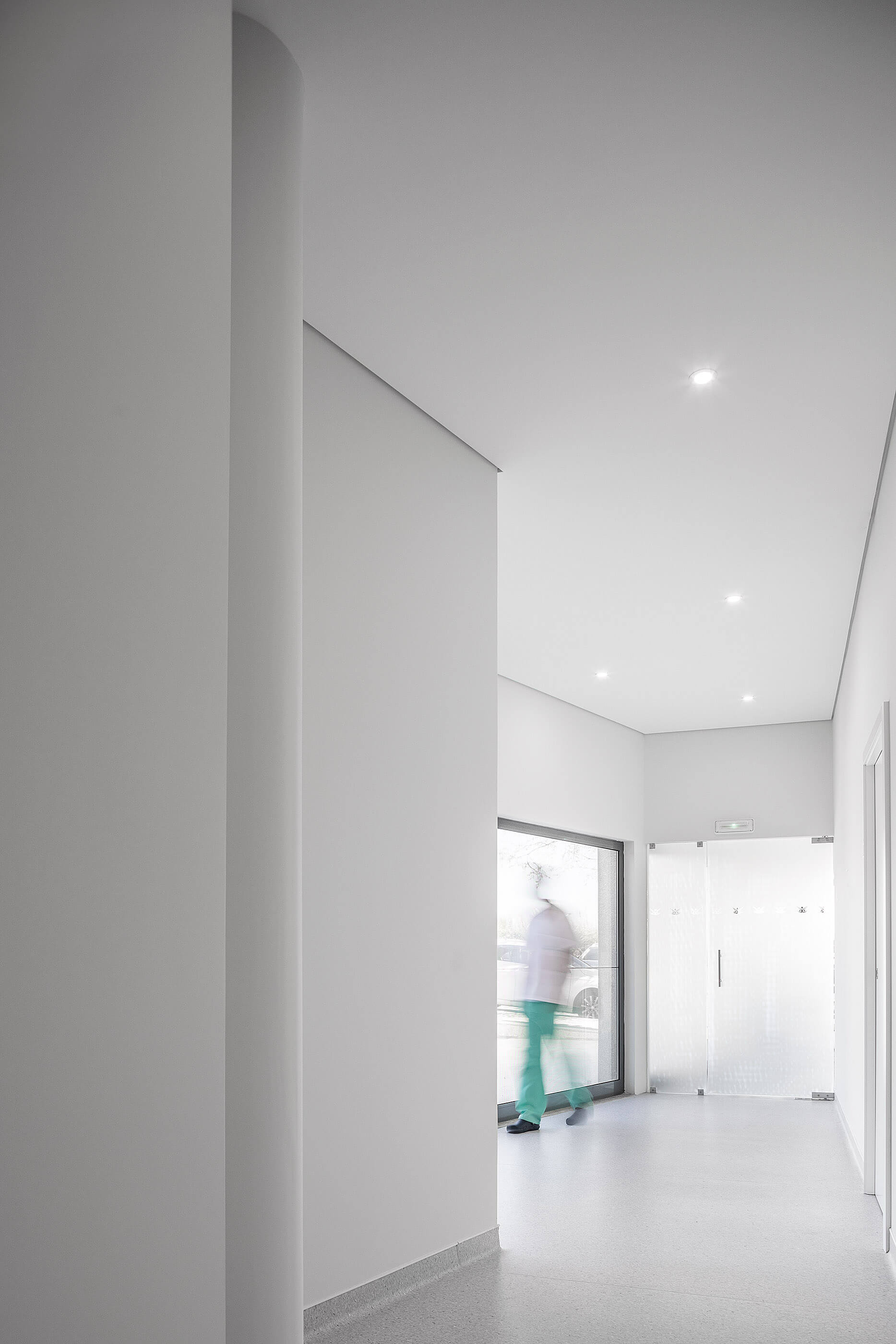 Corredor branco com pavimento cinza, iluminação embutida e porta de vidro ao fundo; pessoa desfocada. — Clínica de Fisioterapia Mar Saúde arquitetura Hinterland Architecture Studio em Vila Nova de Gaia