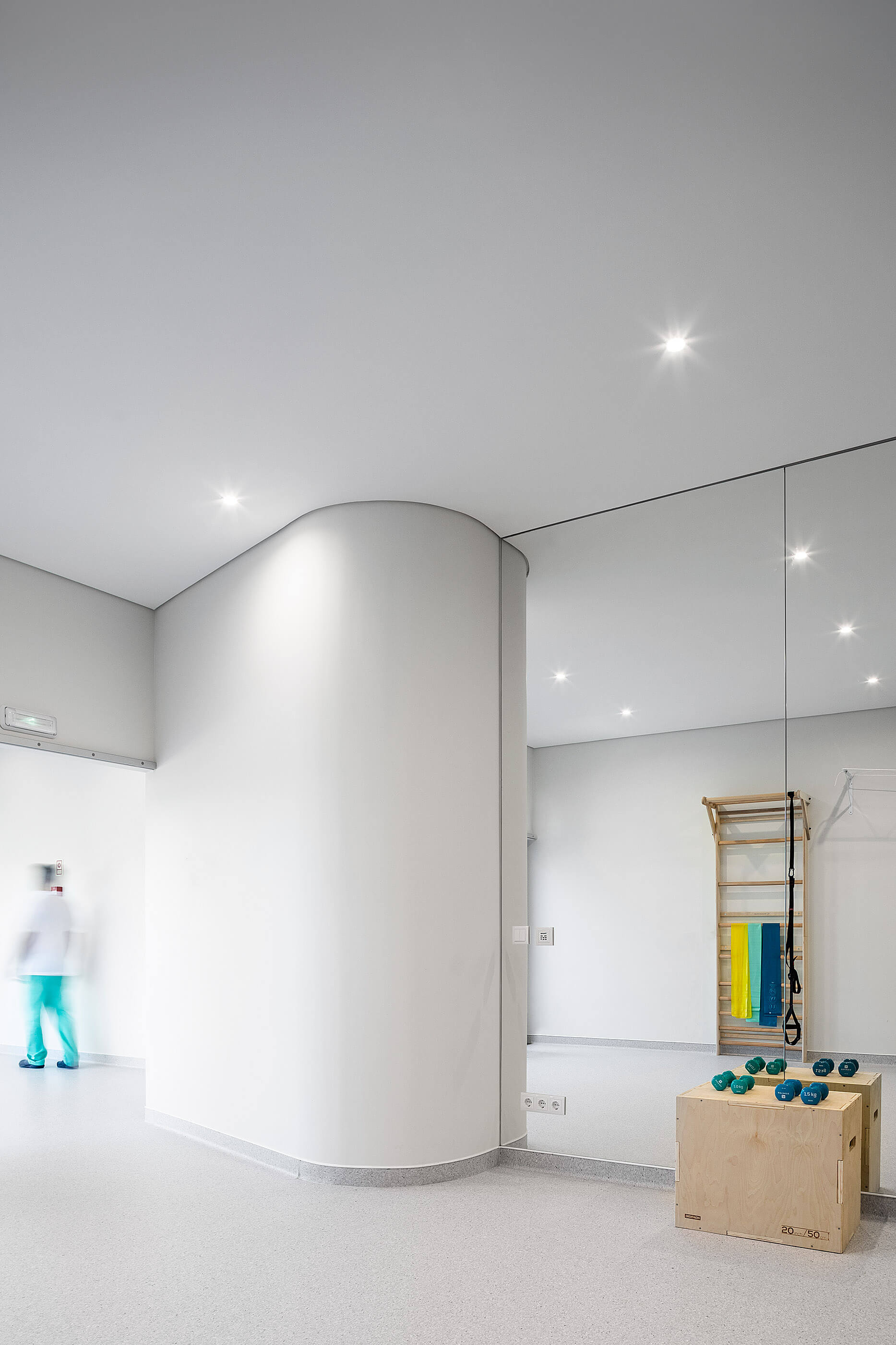 Canto da sala de exercício com espelho, parede curva branca e equipamentos; pavimento cinza. — Clínica de Fisioterapia Mar Saúde arquitetura Hinterland Architecture Studio em Vila Nova de Gaia