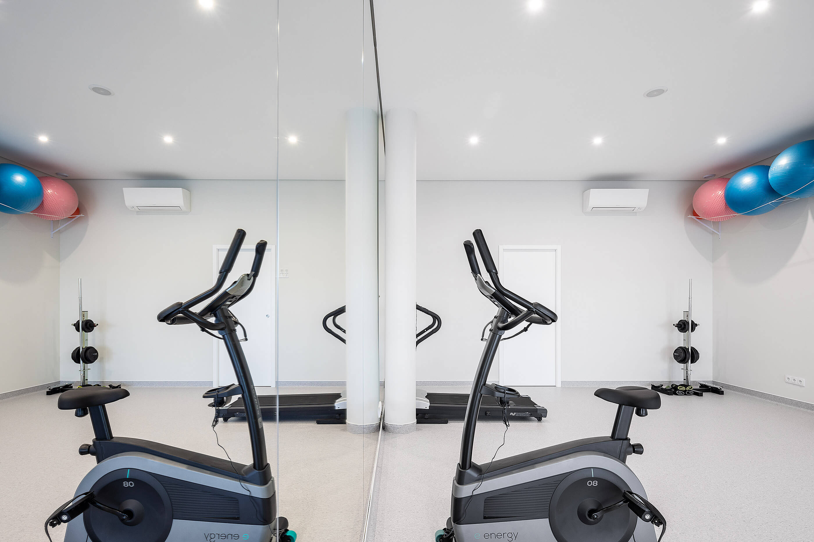 Reflexo no espelho da sala de exercício, com bicicletas estáticas, halteres e bolas de ginástica. — Clínica de Fisioterapia Mar Saúde arquitetura Hinterland Architecture Studio em Vila Nova de Gaia