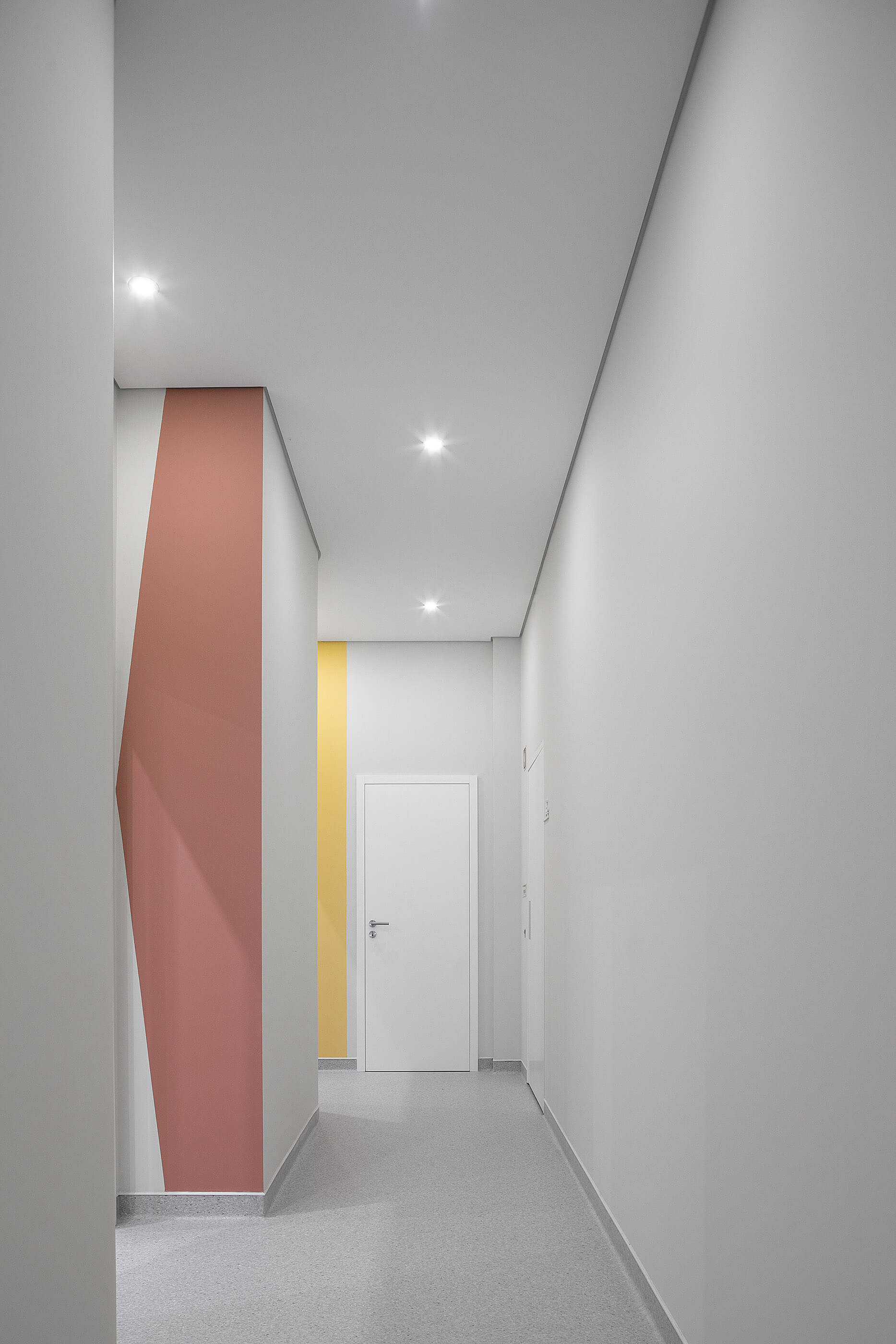 Corredor com paredes brancas e detalhes coloridos em rosa e amarelo; pavimento cinza e iluminação embutida. — Clínica de Fisioterapia Mar Saúde arquitetura Hinterland Architecture Studio em Vila Nova de Gaia