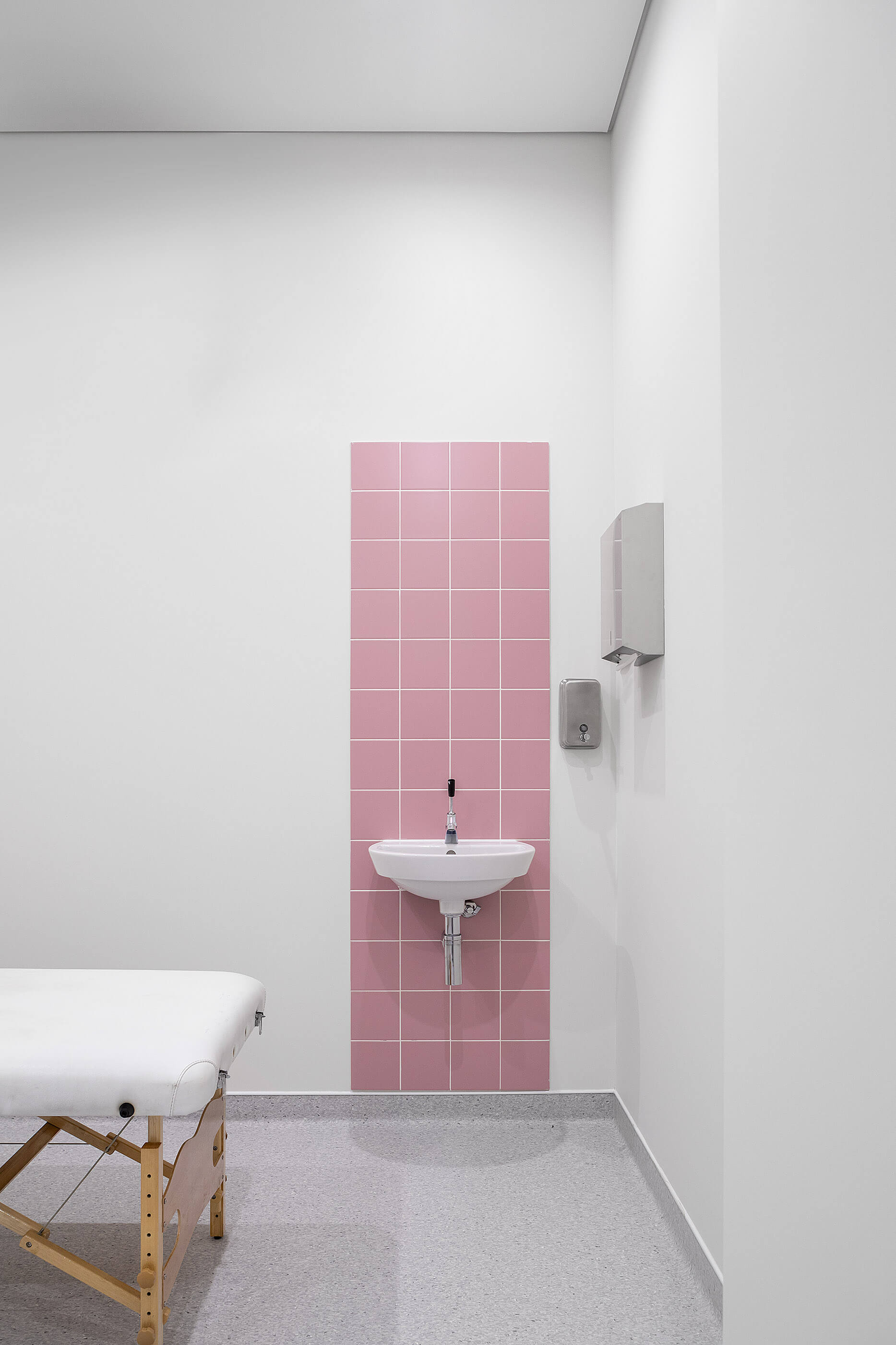 Sala de tratamento com marquesa branca, lavatório com azulejos rosa e pavimento cinza; ambiente clean. — Clínica de Fisioterapia Mar Saúde arquitetura Hinterland Architecture Studio em Vila Nova de Gaia