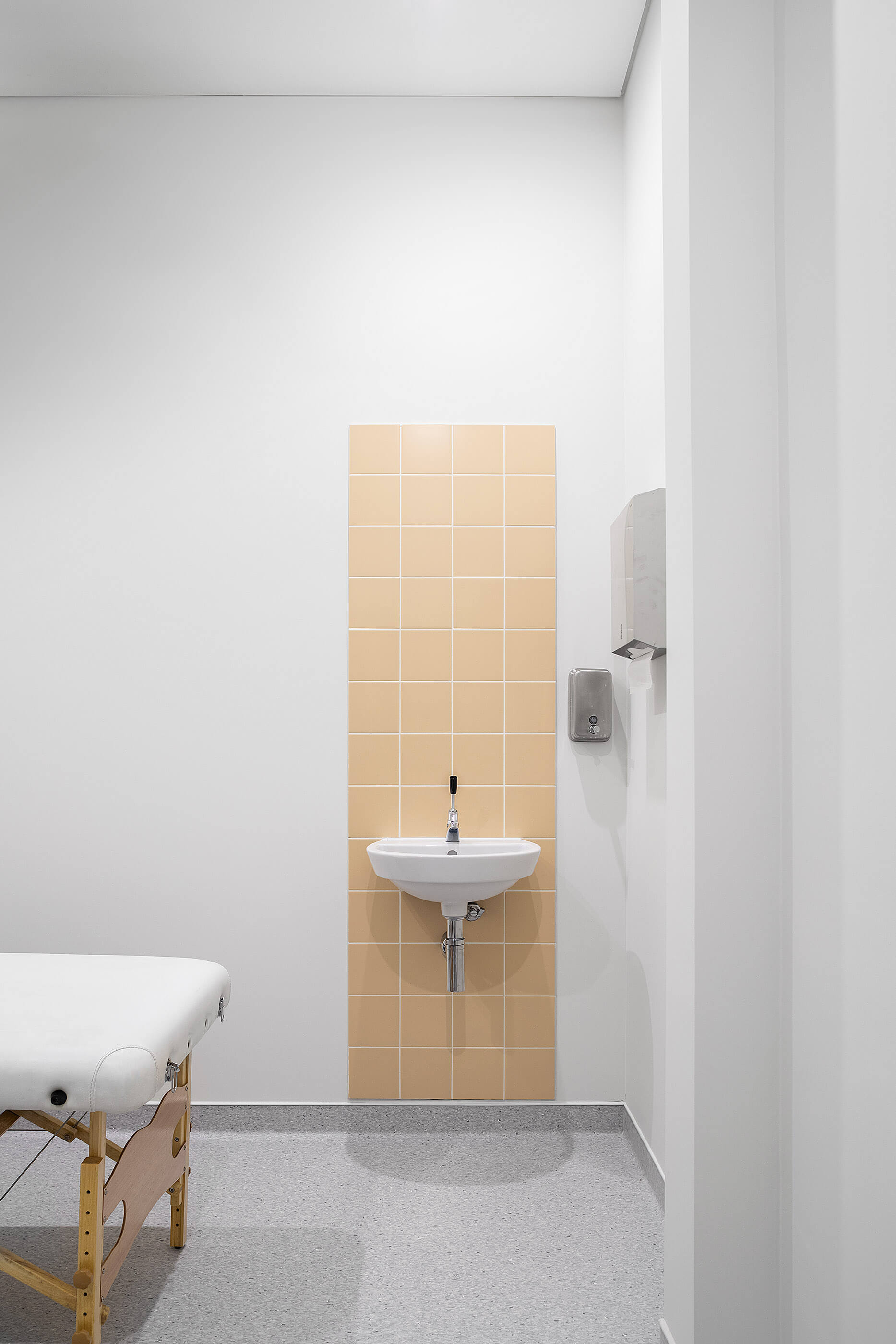 Sala de tratamento com marquesa branca, lavatório com azulejos bege e pavimento cinza; ambiente luminoso. — Clínica de Fisioterapia Mar Saúde arquitetura Hinterland Architecture Studio em Vila Nova de Gaia