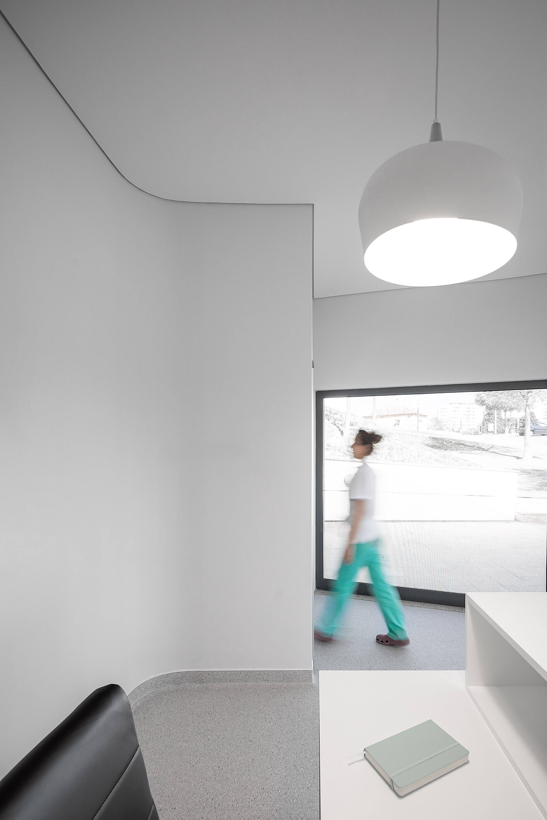 Detalhe da receção com balcão branco, candeeiro suspenso e pavimento cinza; pessoa desfocada ao fundo. — Clínica de Fisioterapia Mar Saúde arquitetura Hinterland Architecture Studio em Vila Nova de Gaia