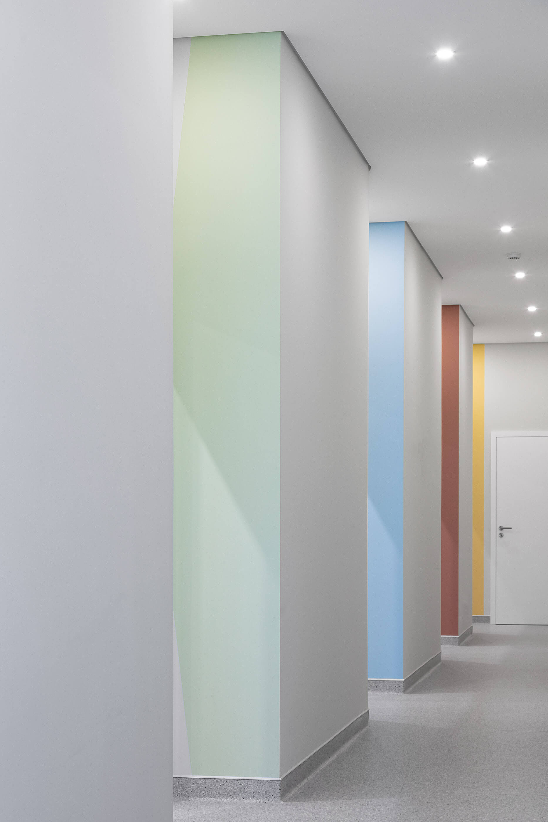 Corredor com paredes brancas e apontamentos coloridos em verde, azul e amarelo; pavimento cinza. — Clínica de Fisioterapia Mar Saúde arquitetura Hinterland Architecture Studio em Vila Nova de Gaia