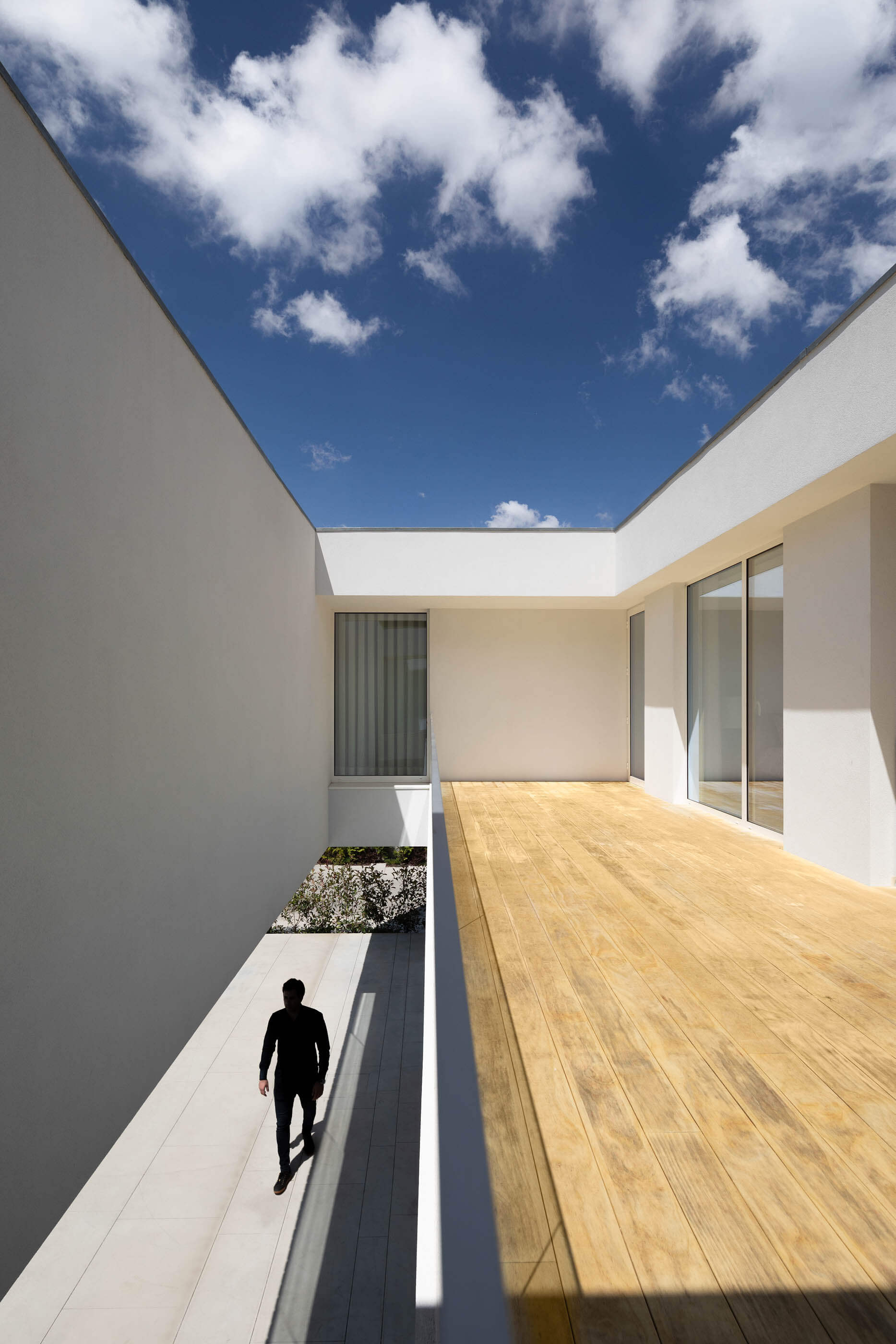 Pátio interior branco com pavimento em madeira e pedra, sob um céu azul com nuvens. — Casa A arquitetura REM'A Arquitectos em Portugal