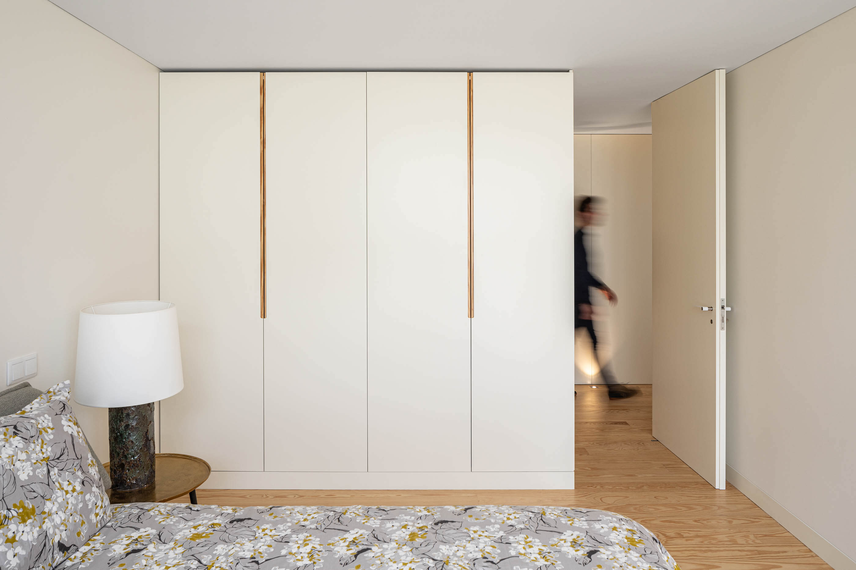 Quarto com armário branco de portas altas, cama com roupa de cama estampada e chão de madeira. — Casa A arquitetura REM'A Arquitectos em Portugal