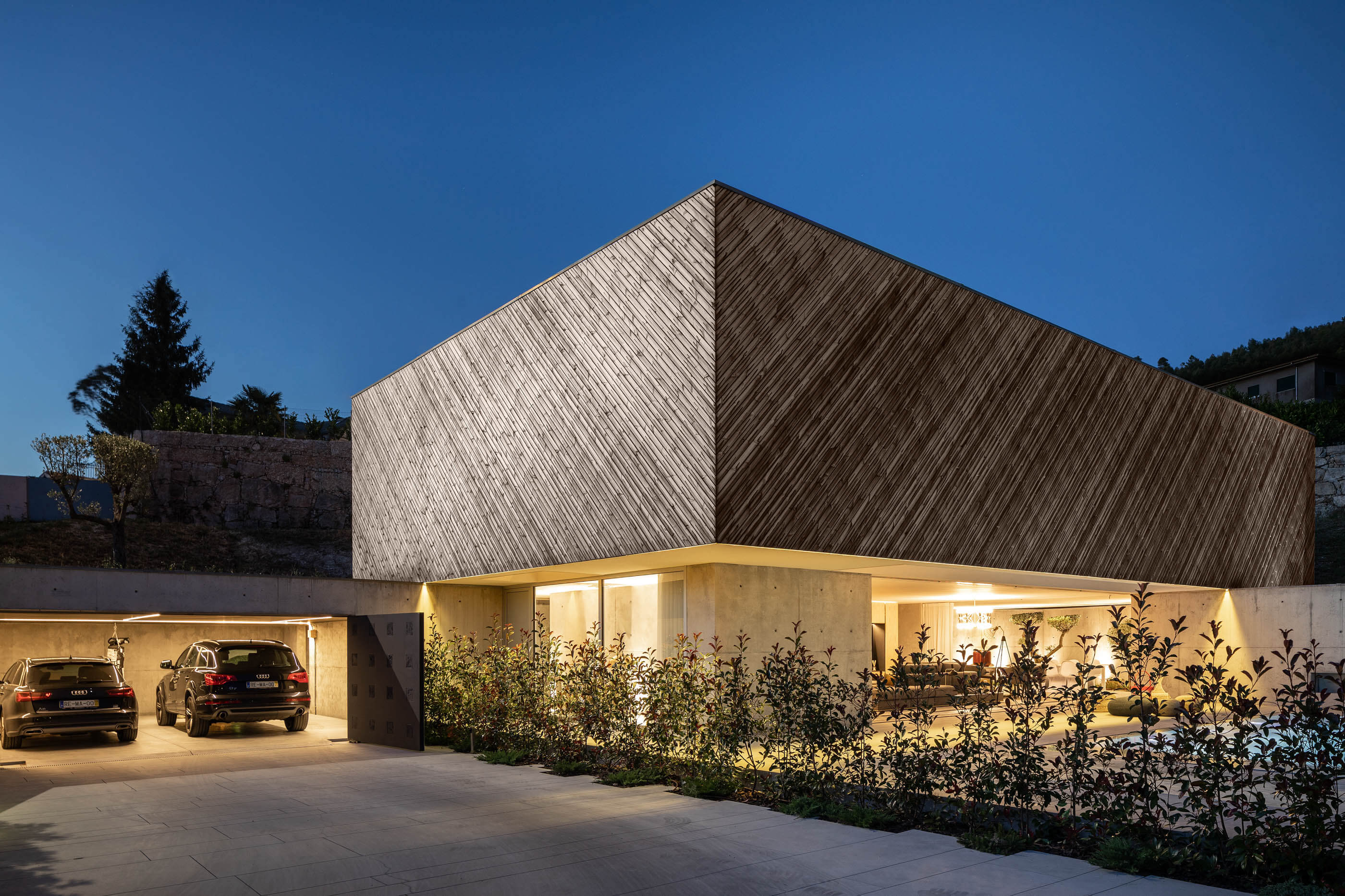 Vista noturna da Casa A com garagem, fachada em madeira e iluminação exterior. — Casa A arquitetura REM'A Arquitectos em Portugal