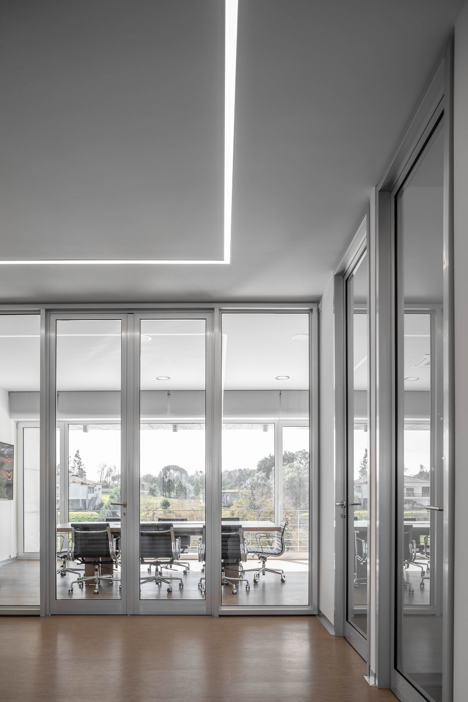 Corredor com divisórias de vidro que revelam salas de reunião, pavimento em madeira clara. — Edifício Sede PRF arquitetura Impare Arquitectura em Portugal