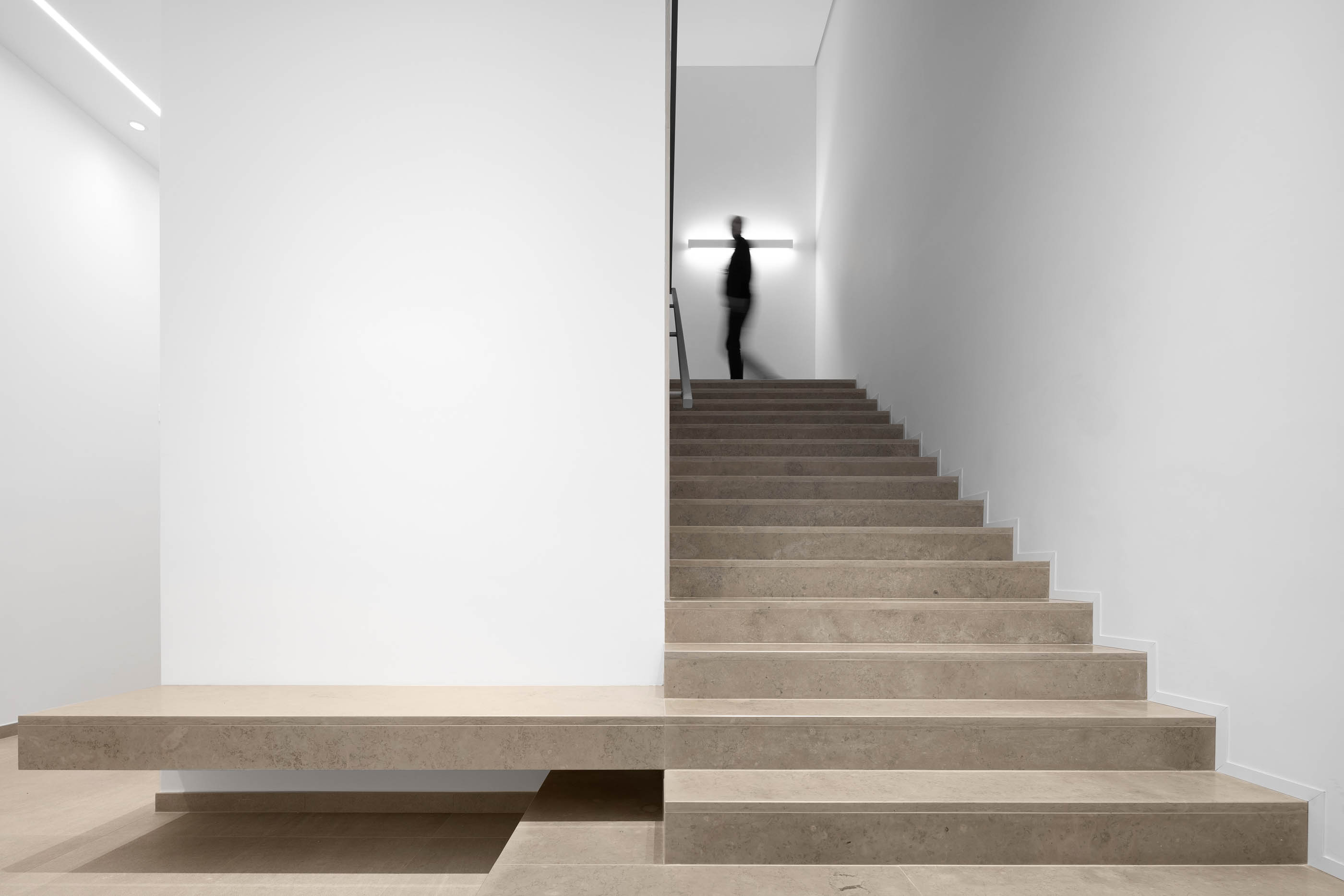Escadaria em pedra clara com paredes brancas, iluminação indireta e silhueta de pessoa. — Edifício Sede PRF arquitetura Impare Arquitectura em Portugal