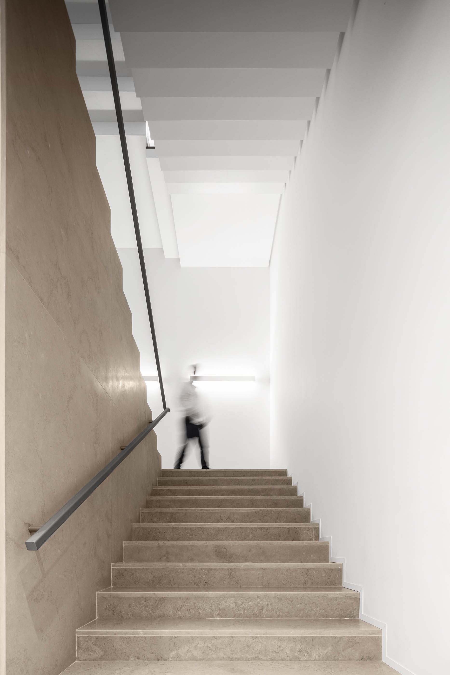 Escadaria com revestimento em pedra clara e parede com textura, iluminação linear. — Edifício Sede PRF arquitetura Impare Arquitectura em Portugal
