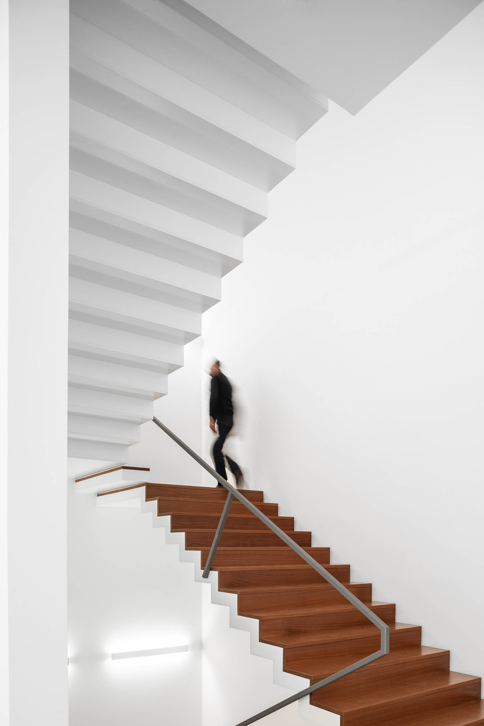 Escadaria com degraus em madeira, estrutura branca e iluminação indireta, pessoa a subir. — Edifício Sede PRF arquitetura Impare Arquitectura em Portugal