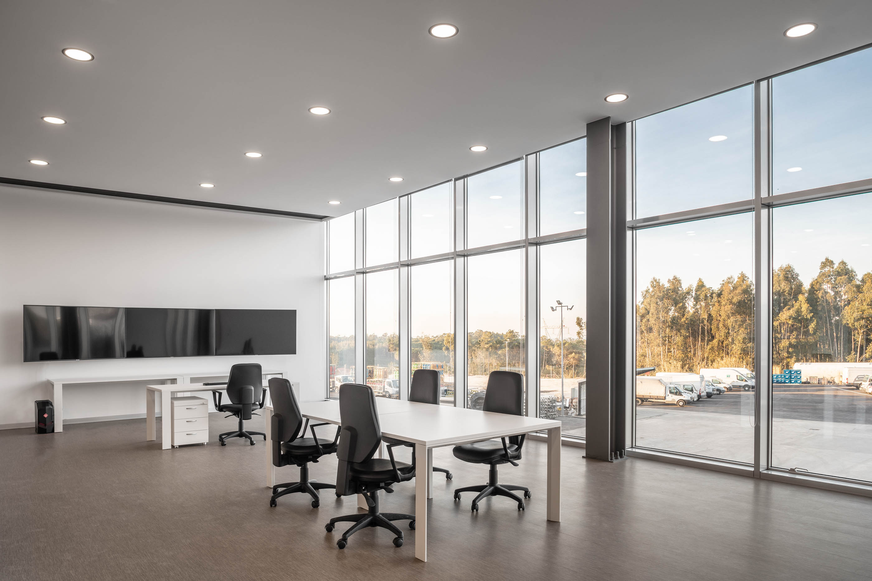 Espaço amplo com mesas de trabalho, cadeiras pretas e janelas panorâmicas com vista exterior. — Edifício Sede PRF arquitetura Impare Arquitectura em Portugal