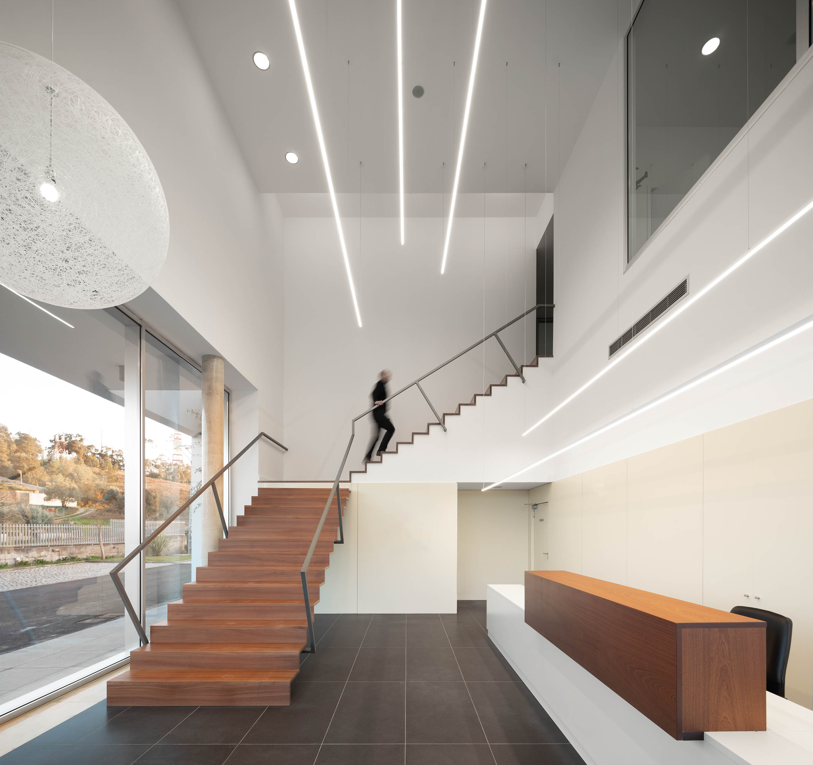 Hall de entrada com escadaria em madeira, iluminação linear e candeeiro esférico suspenso. — Edifício Sede PRF arquitetura Impare Arquitectura em Portugal