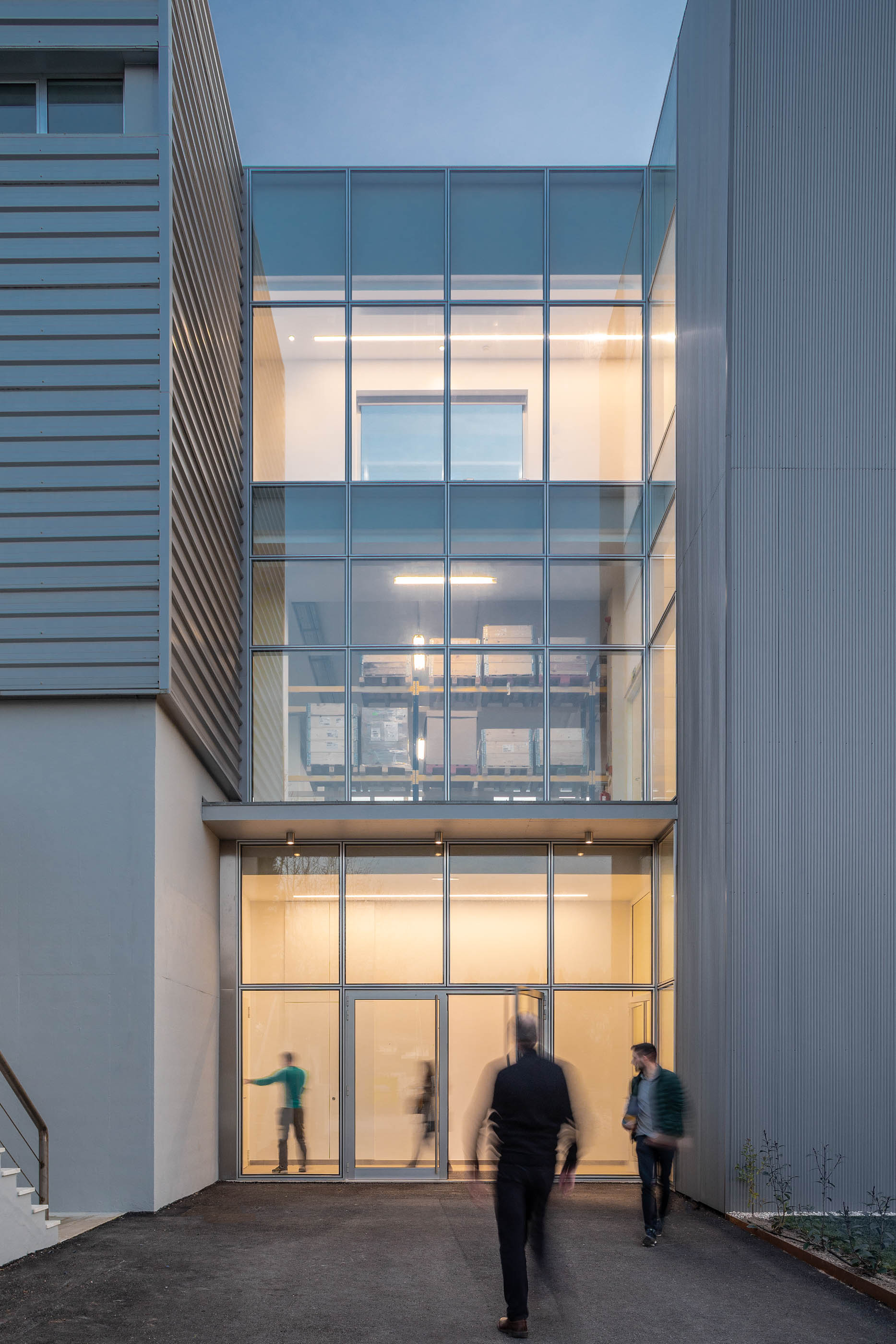 Fachada envidraçada com estrutura metálica, pessoas a entrar no edifício, iluminação interior. — Edifício Sede PRF arquitetura Impare Arquitectura em Portugal