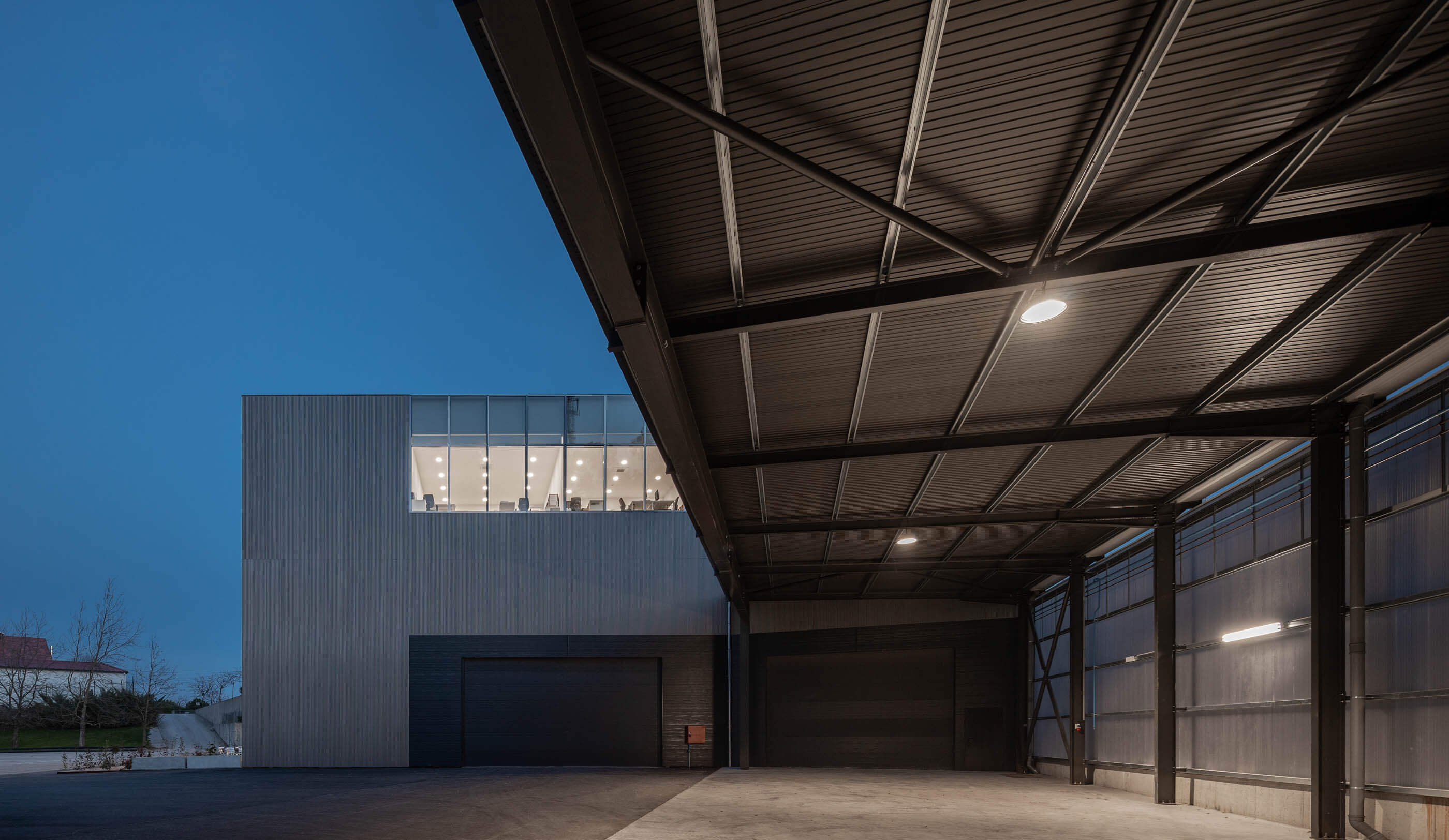 Detalhe noturno de armazém com estrutura metálica, iluminação e edifício ao fundo. — Edifício Sede PRF arquitetura Impare Arquitectura em Portugal