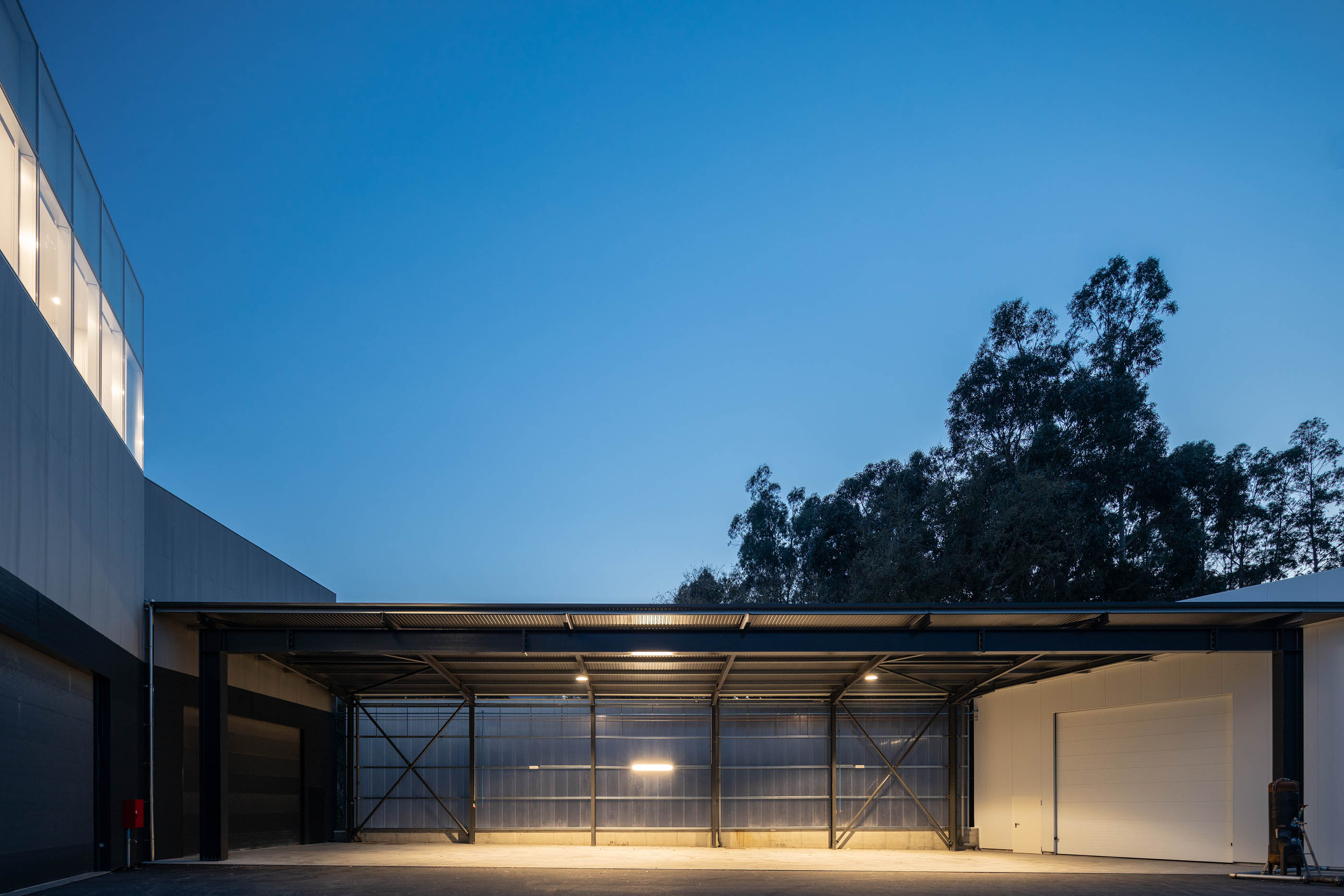 Estrutura metálica de armazém com iluminação noturna, céu azul escuro e vegetação ao fundo. — Edifício Sede PRF arquitetura Impare Arquitectura em Portugal