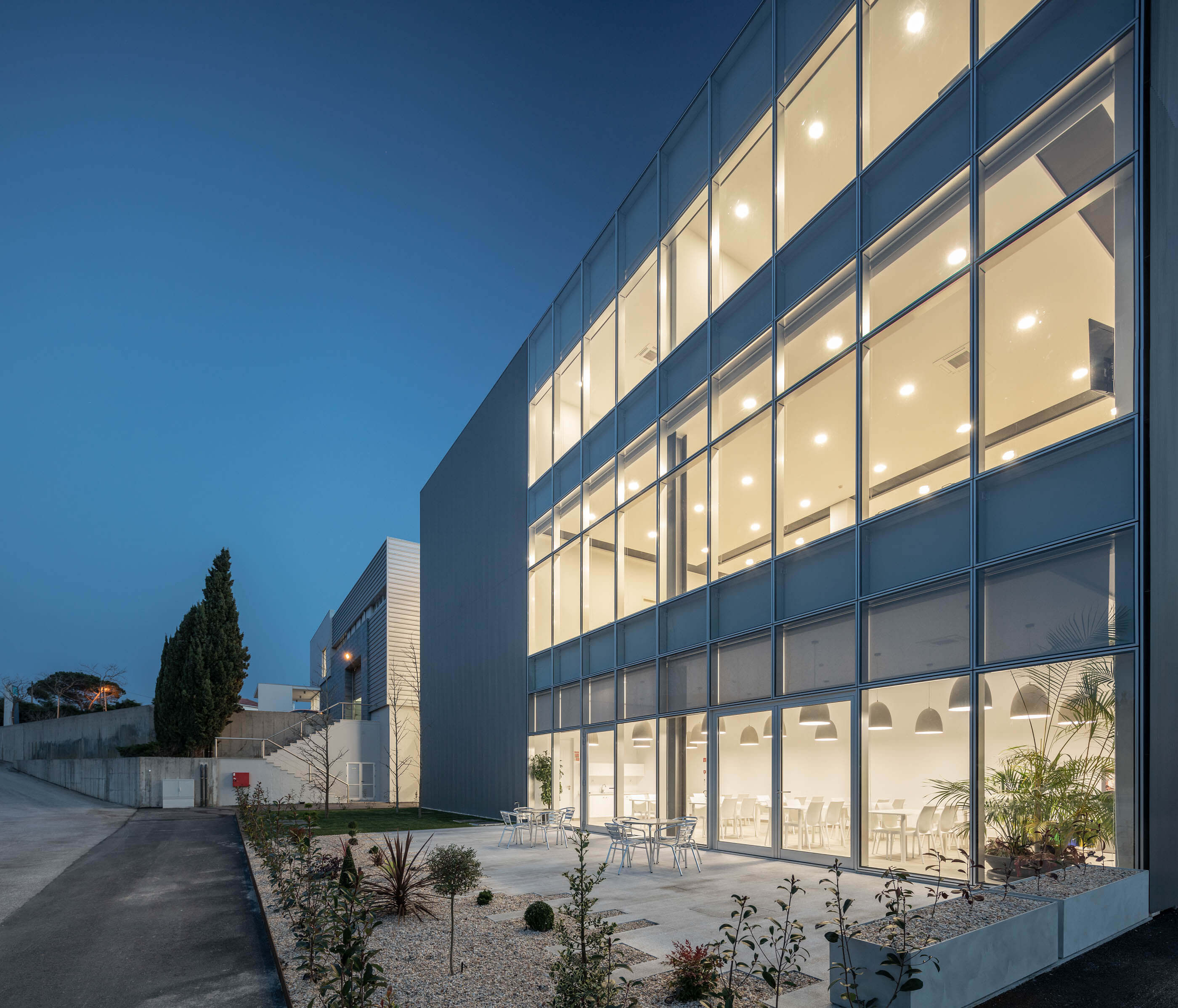 Fachada envidraçada iluminada à noite, com área exterior ajardinada e mobiliário de jardim. — Edifício Sede PRF arquitetura Impare Arquitectura em Portugal