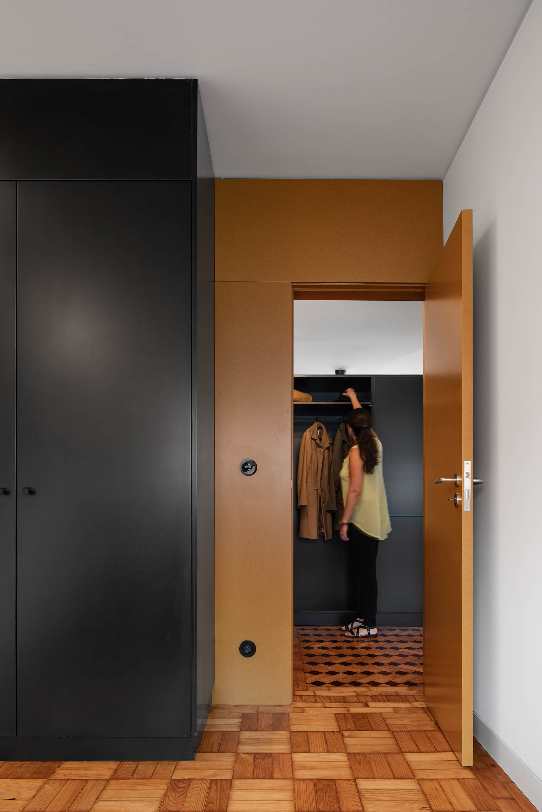 Entrada para closet com armário preto, porta cor de mel e pavimento em madeira com padrão geométrico. — Apartamento Morro arquitetura Hinterland Architecture Studio em Vila Nova de Gaia