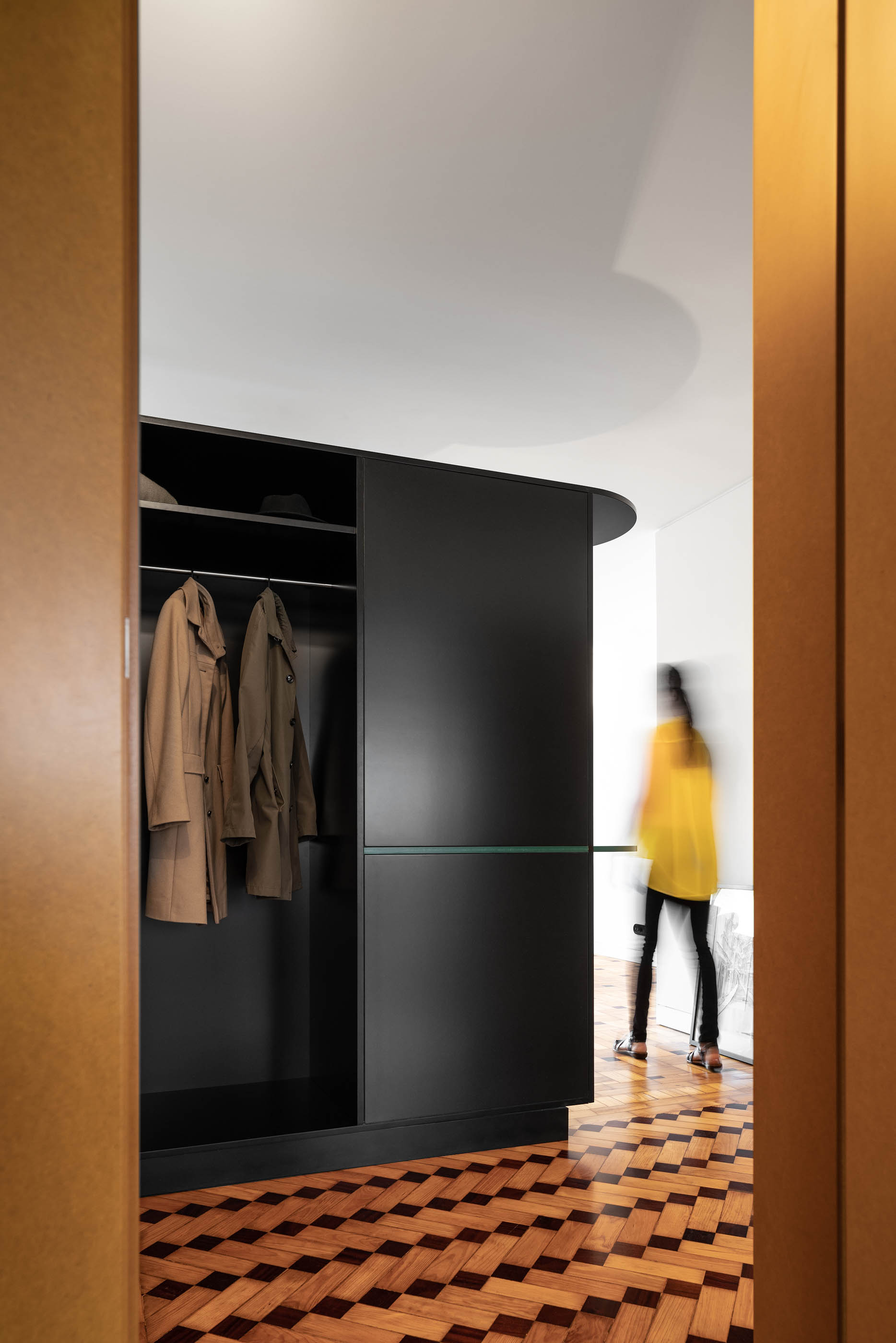 Interior de closet com armário preto curvo e pavimento em madeira com padrão em espinha. — Apartamento Morro arquitetura Hinterland Architecture Studio em Vila Nova de Gaia