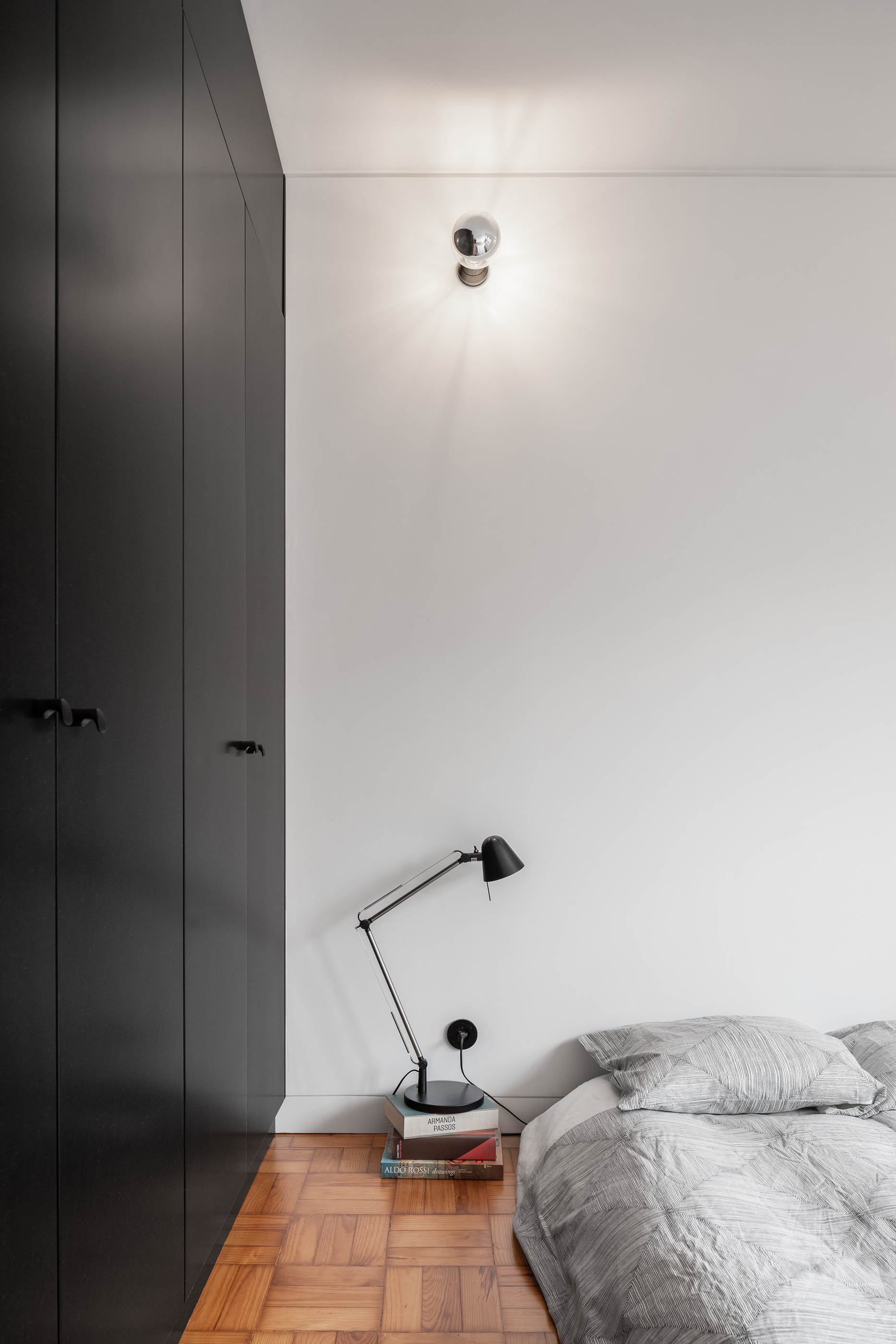 Quarto com cama, armário preto, candeeiro de mesa e aplique de parede, pavimento em madeira. — Apartamento Morro arquitetura Hinterland Architecture Studio em Vila Nova de Gaia