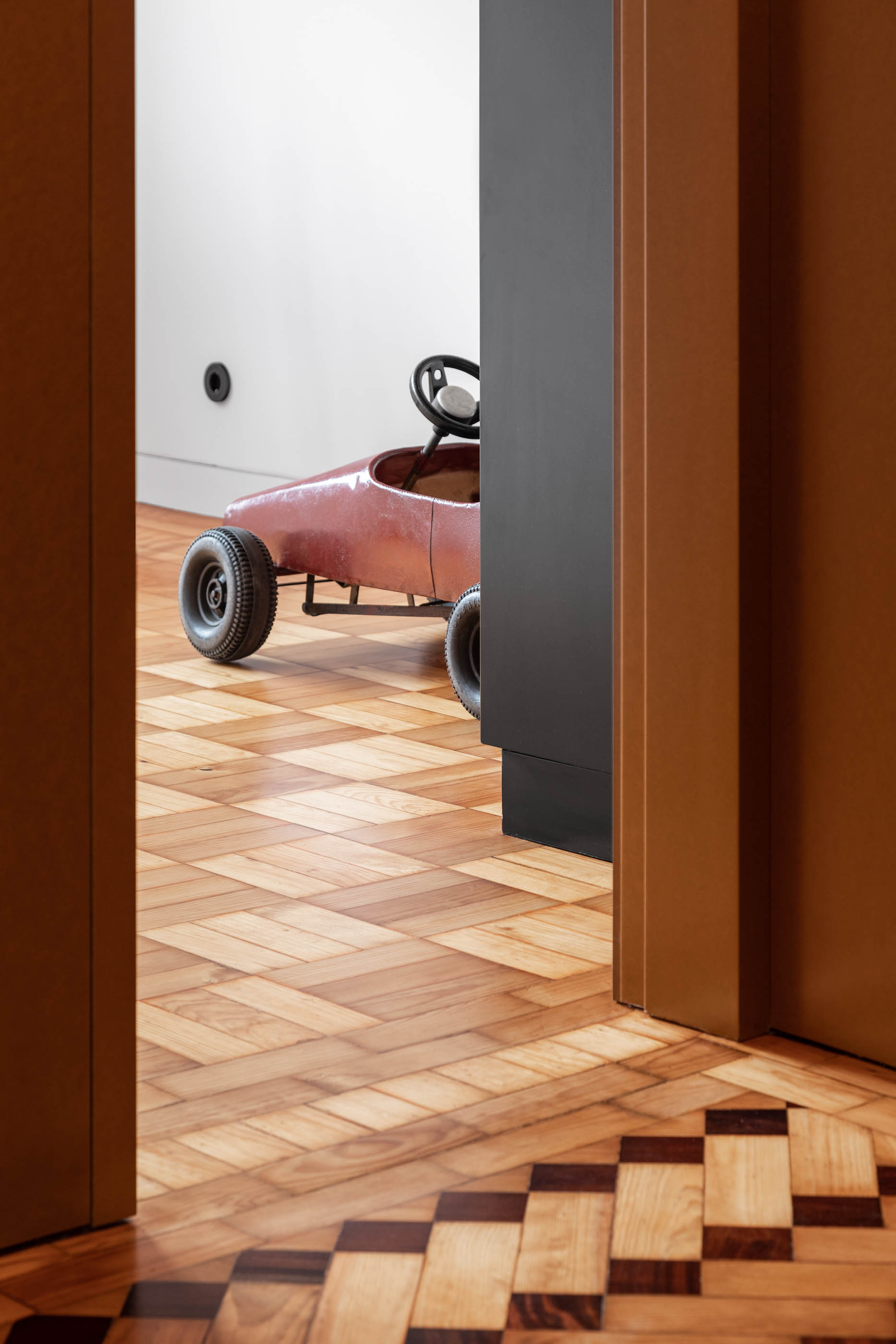 Quarto com carro de brincar vermelho, pavimento em madeira com padrão geométrico e porta em madeira. — Apartamento Morro arquitetura Hinterland Architecture Studio em Vila Nova de Gaia