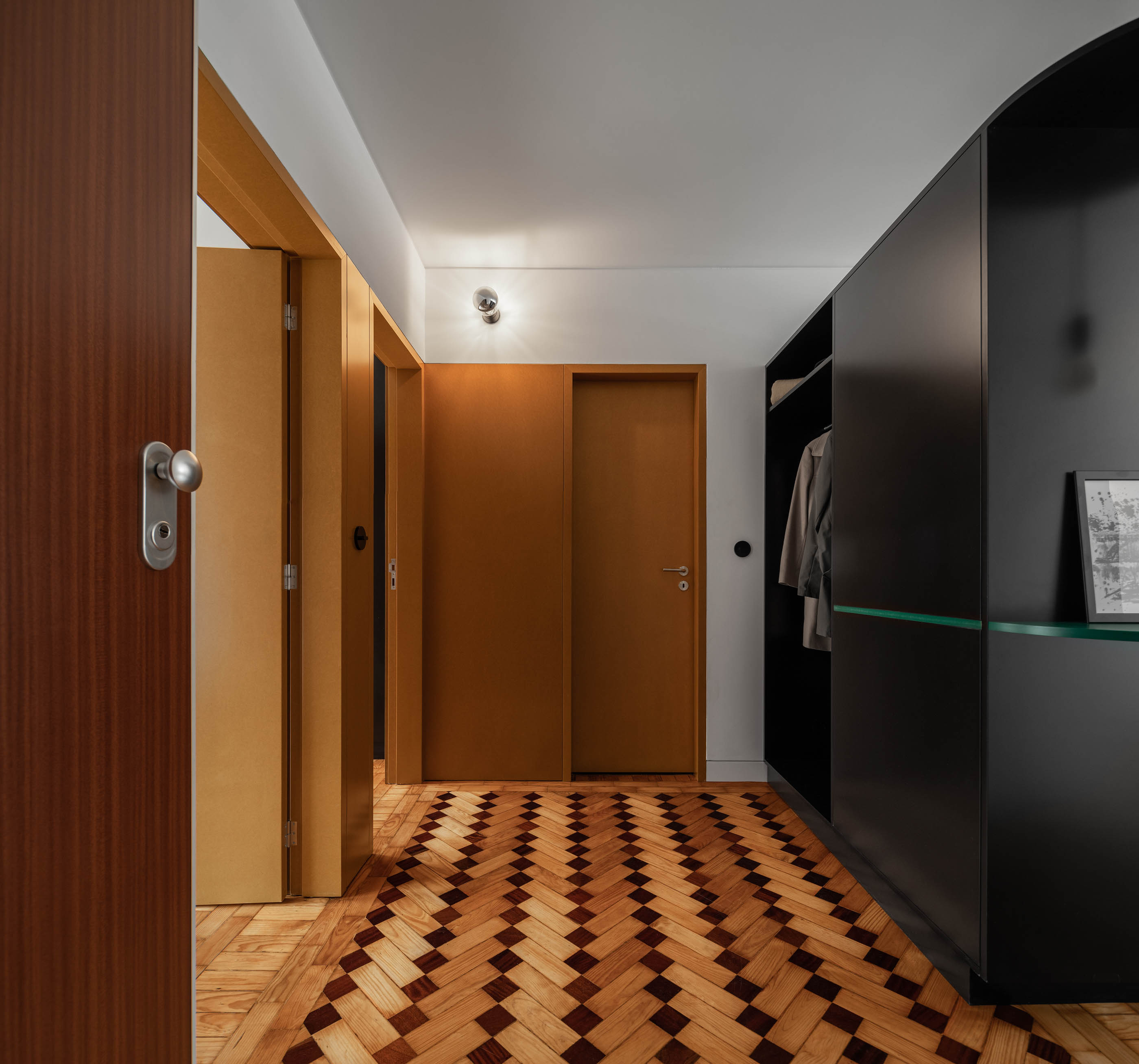 Corredor com portas em madeira cor de mel, armário preto e pavimento em madeira com padrão geométrico. — Apartamento Morro arquitetura Hinterland Architecture Studio em Vila Nova de Gaia