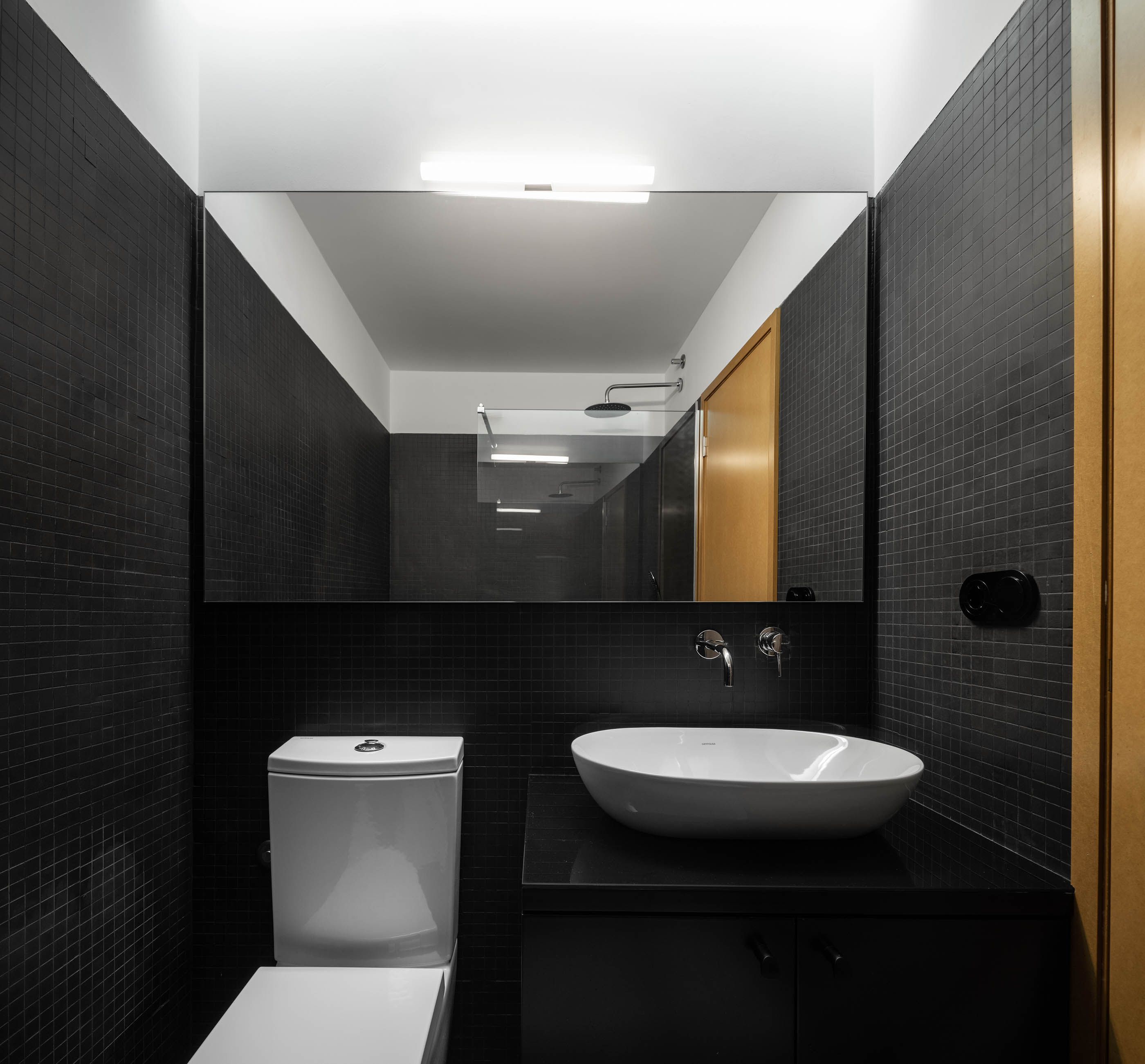 Casa de banho com azulejos pretos, espelho amplo, lavatório branco e porta em madeira cor de mel. — Apartamento Morro arquitetura Hinterland Architecture Studio em Vila Nova de Gaia