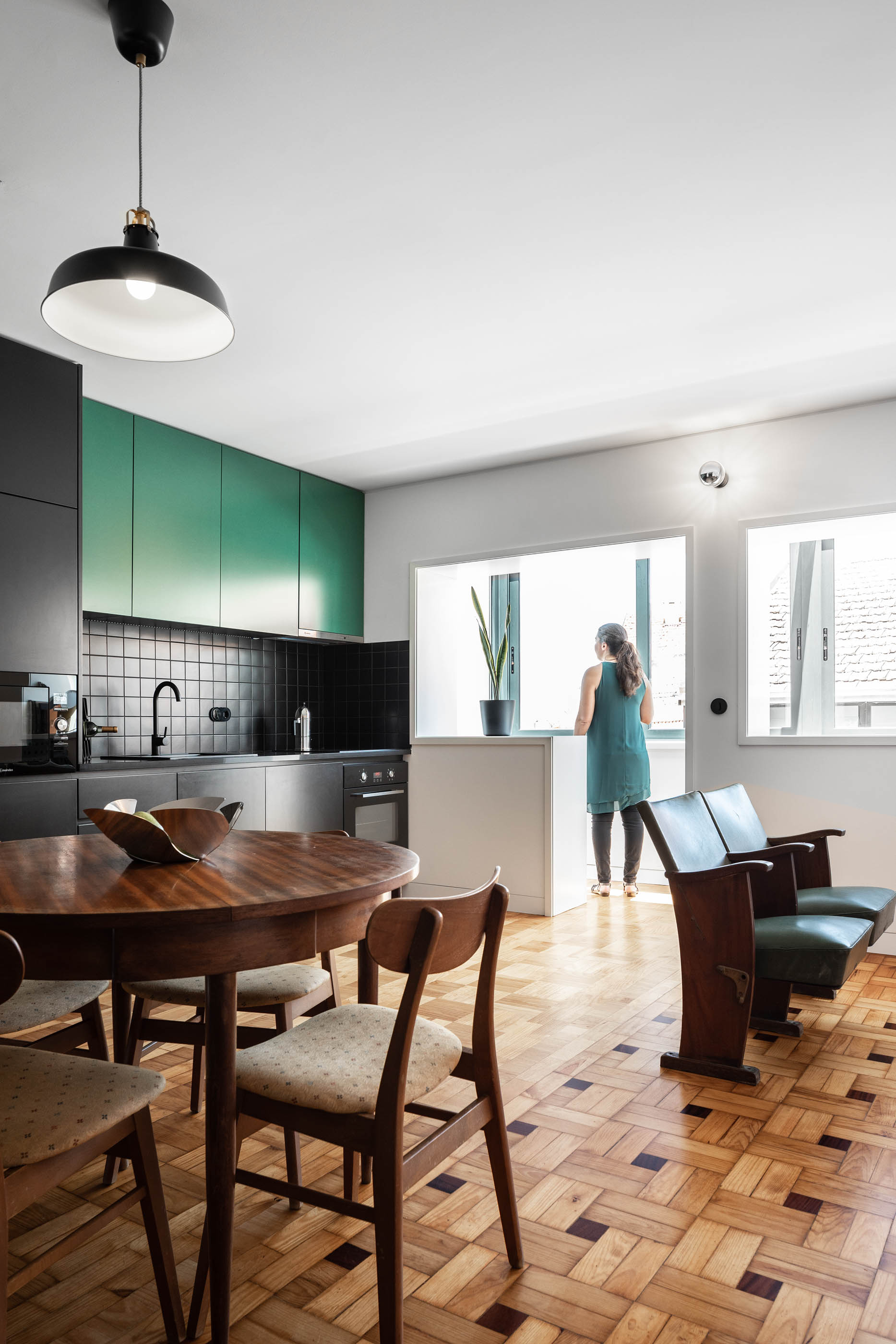 Cozinha com mesa de jantar redonda, cadeiras de madeira e mulher a olhar pela janela. — Apartamento Morro arquitetura Hinterland Architecture Studio em Vila Nova de Gaia